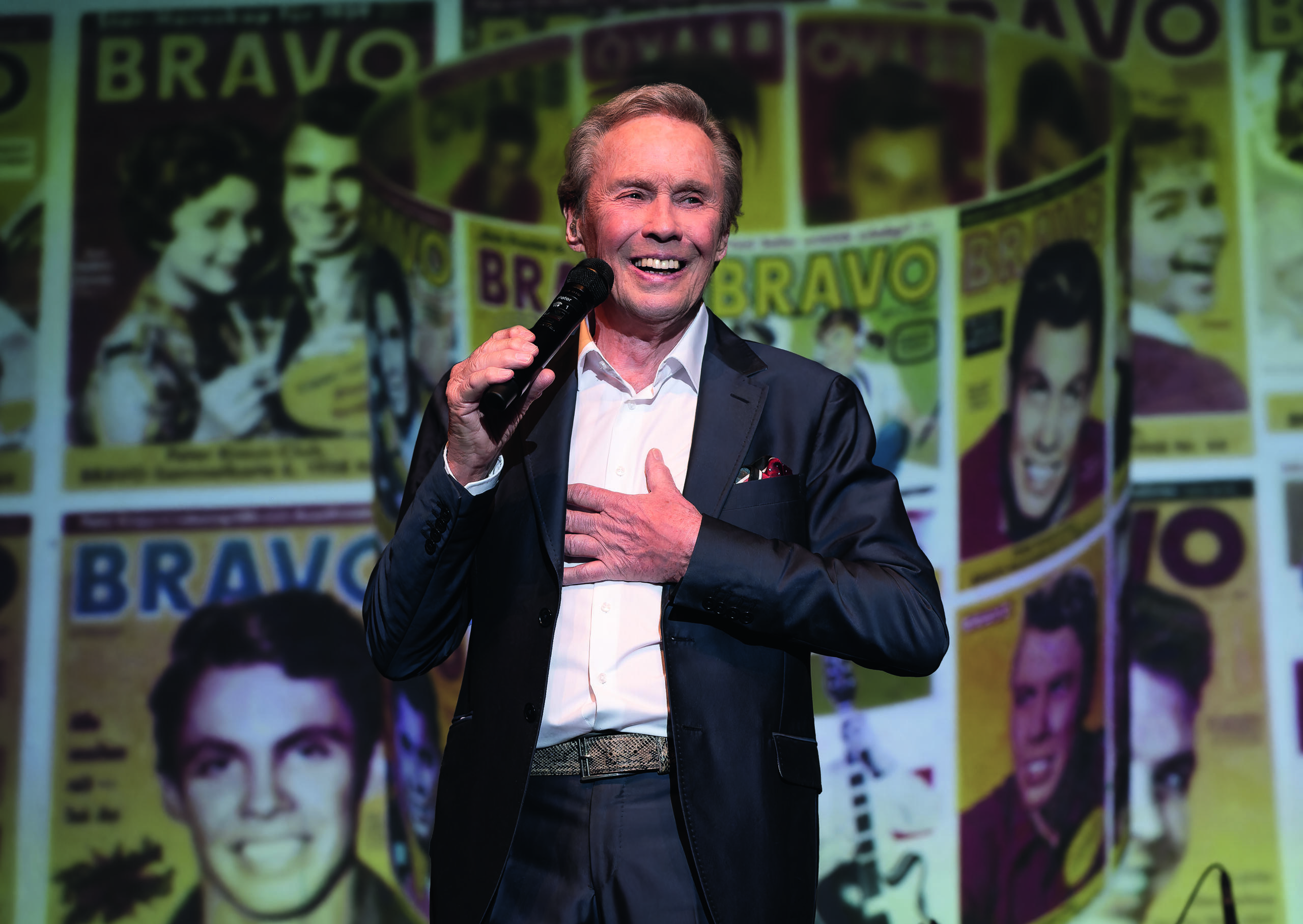 Der Sänger Peter Kraus steht in einem Anzug mit Mikrofon in der Hand auf einer Bühne. Im Hintergrund sind Projektionen von alten Bravo-Covers zu sehen.