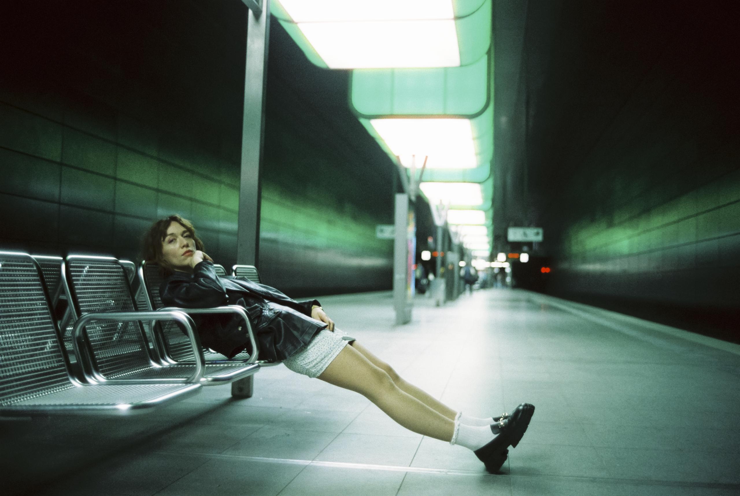 The platform of a subway station. A young woman in a leather jacket and shorts poses on the seats.