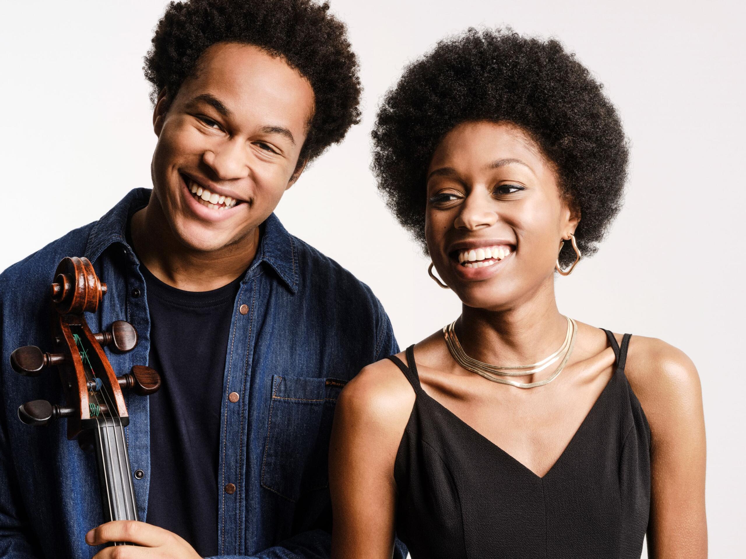 A young man and a young woman close to each other, they both have Afro hairstyles and laugh into the camera.