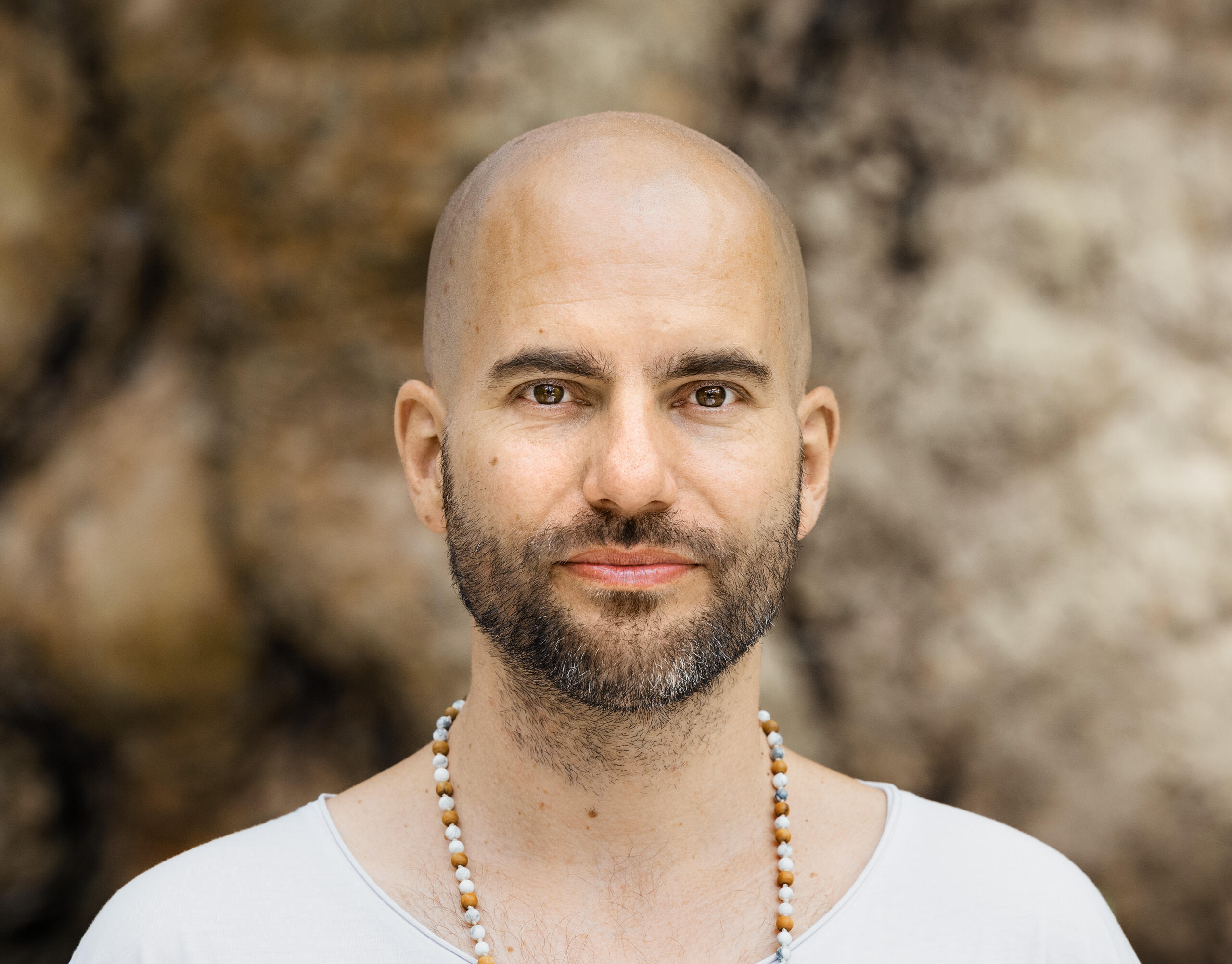 A portrait photograph of the musician Seom. He is wearing a white T-shirt, a necklace of brown and white beads and is looking straight into the camera. Rocks can be seen blurred in the background.