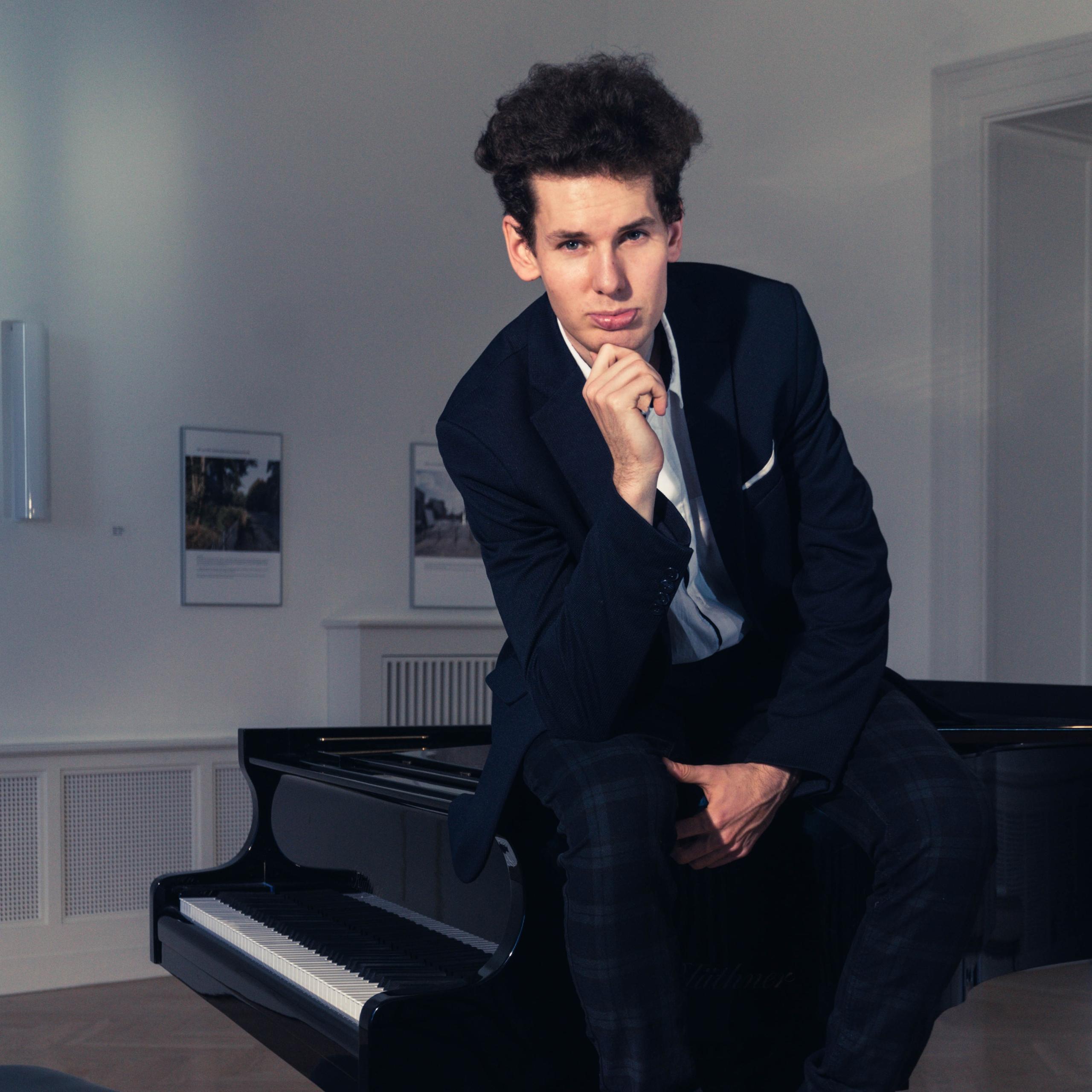 Thomas Krüger sits on his grand piano in a black suit
