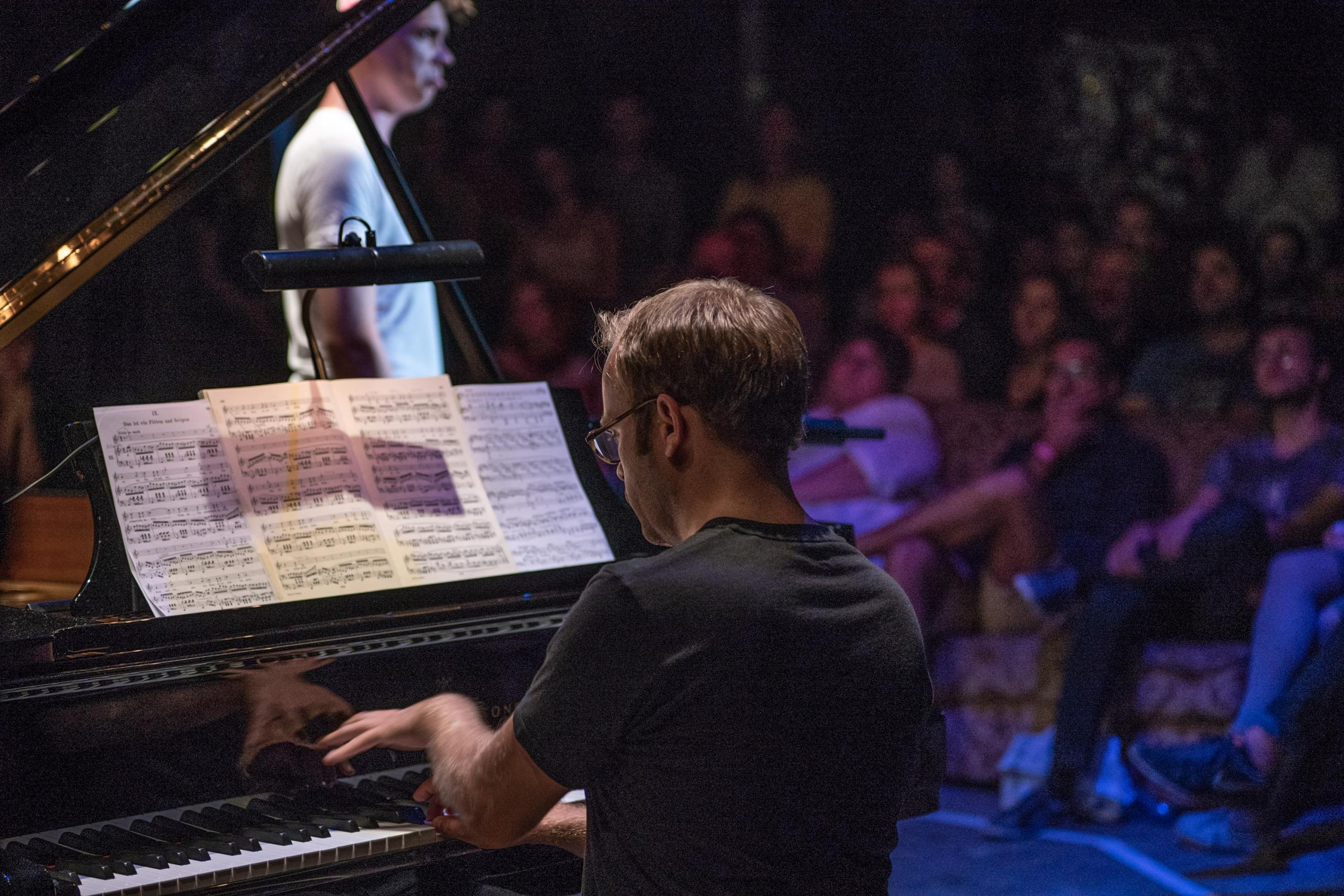 A man plays the grand piano, blurred you can see a singer standing in front of an audience
