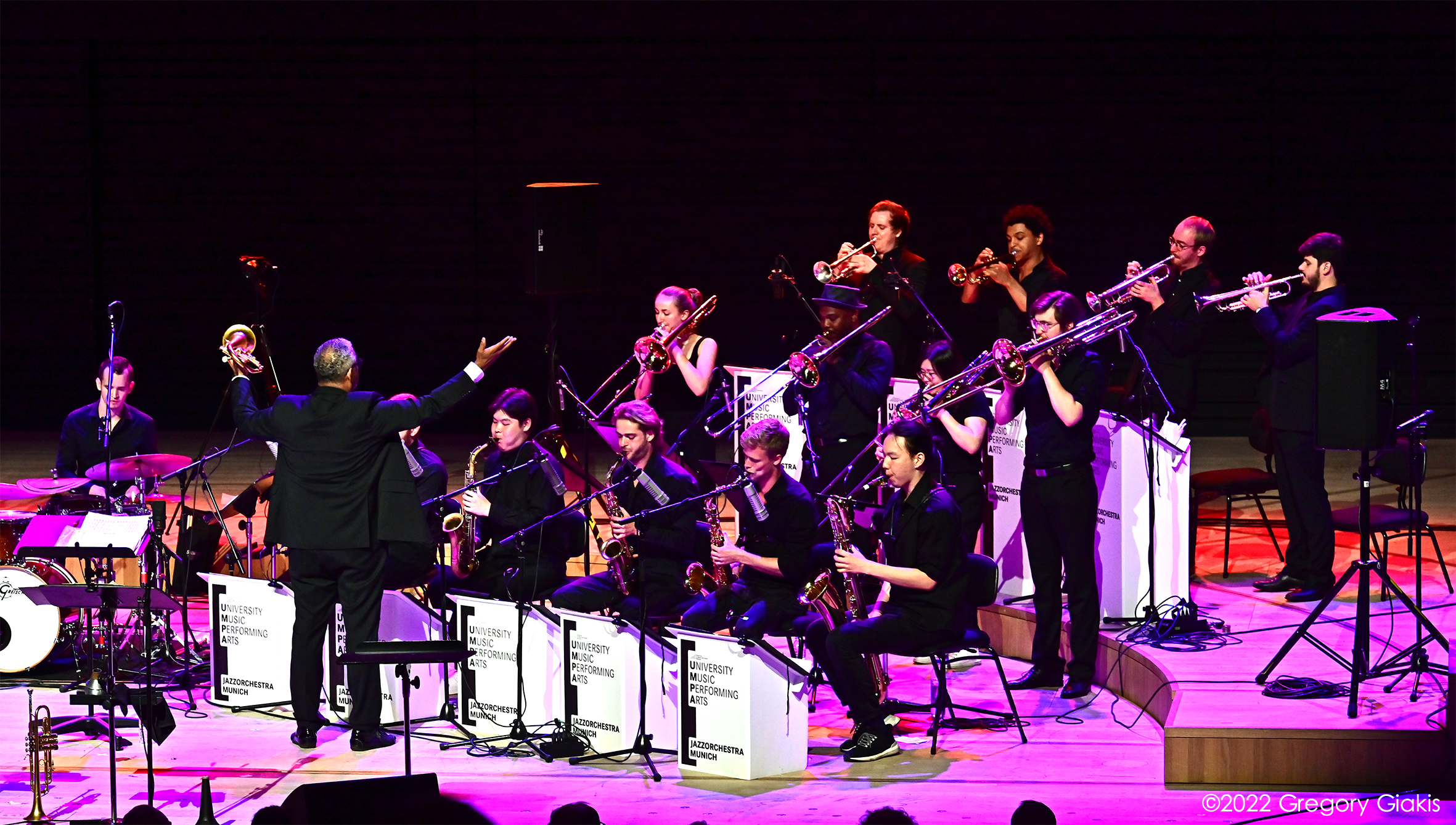 Ein Ensemble von Blechbläsern auf der Bühne, mit dem Rücken zu uns der Dirigent mit erhobenen Armen. 