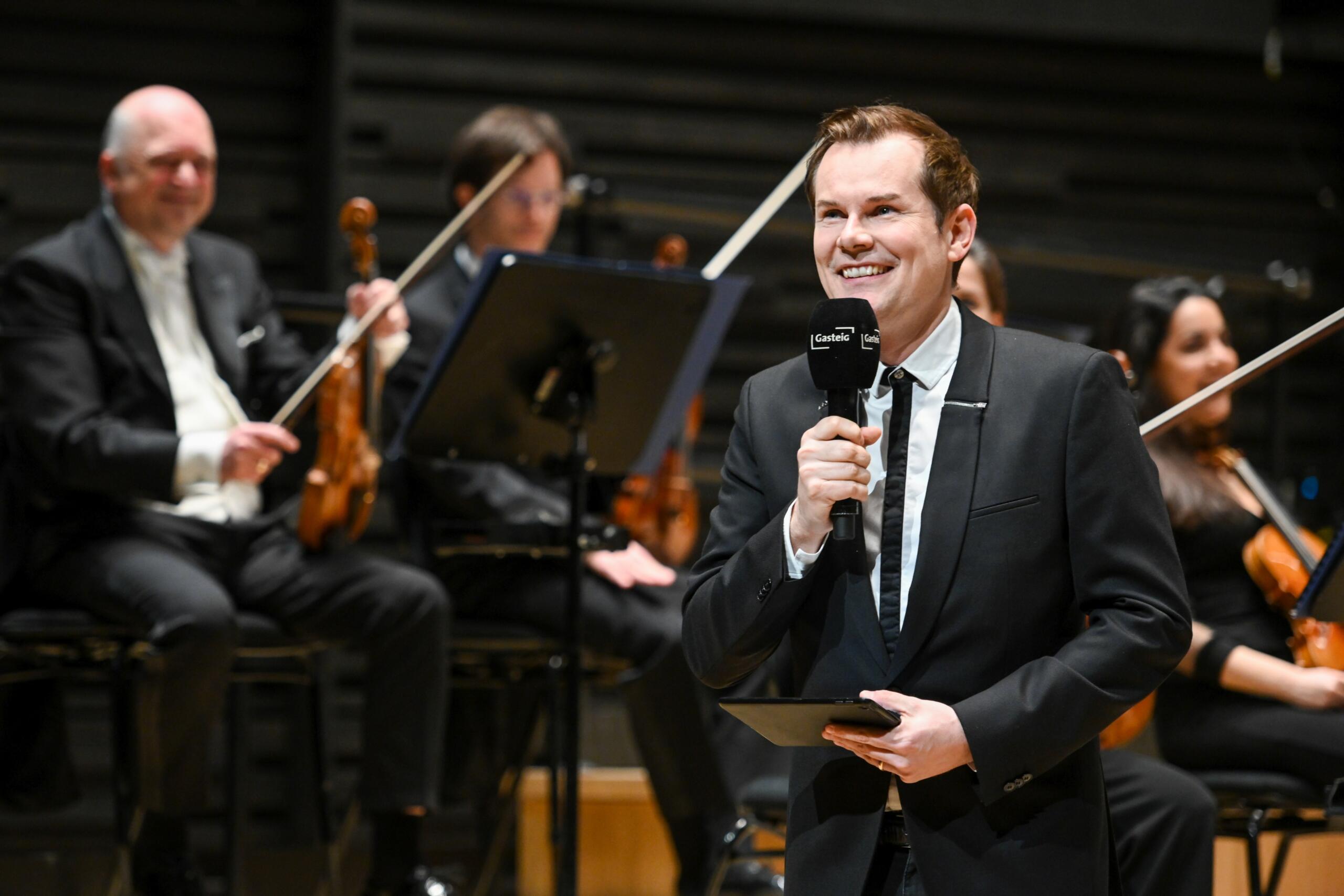 Malte Arkona smiling with microphone on the stage of the Isarphilharmonie. Members of the orchestra can be seen in the background.