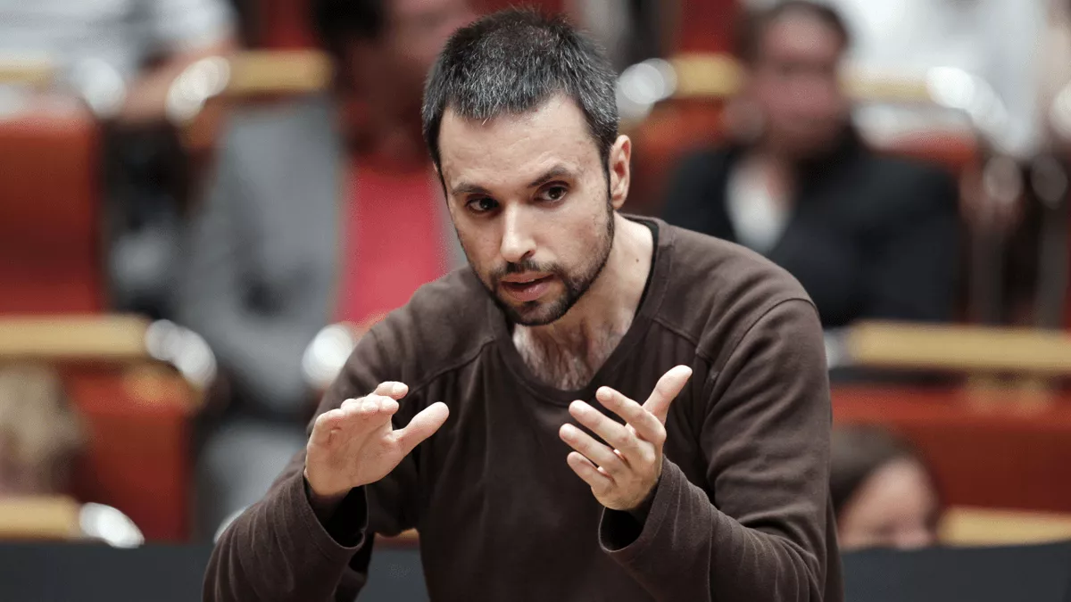 Portrait of the conductor Constantinos Carydis. He is wearing a brown jumper and is depicted conducting.