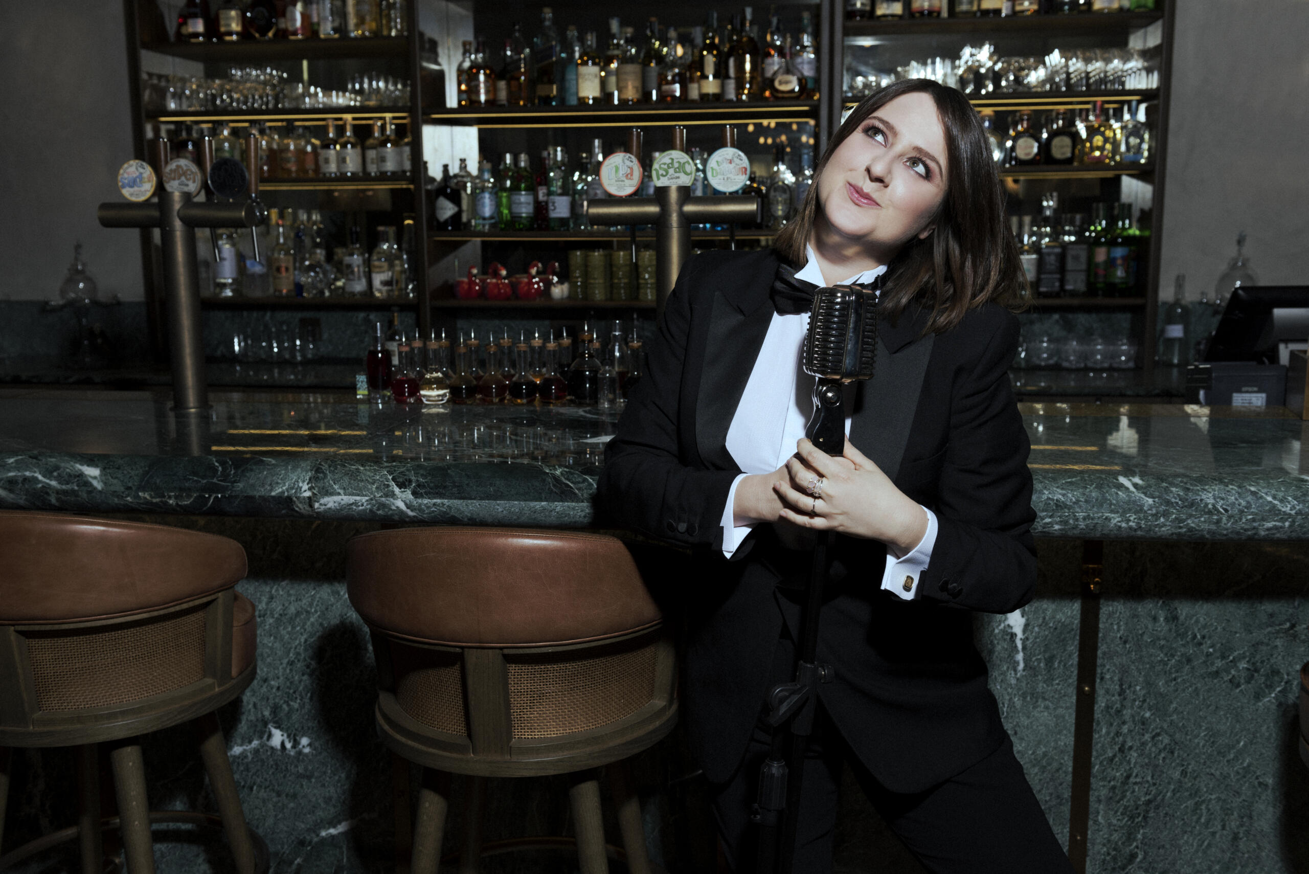 Ein Porträt der italienischen Stand-up-Comedian Michela Giraud. Sie lehnt in einem schwarzen Anzug an einer Bar.