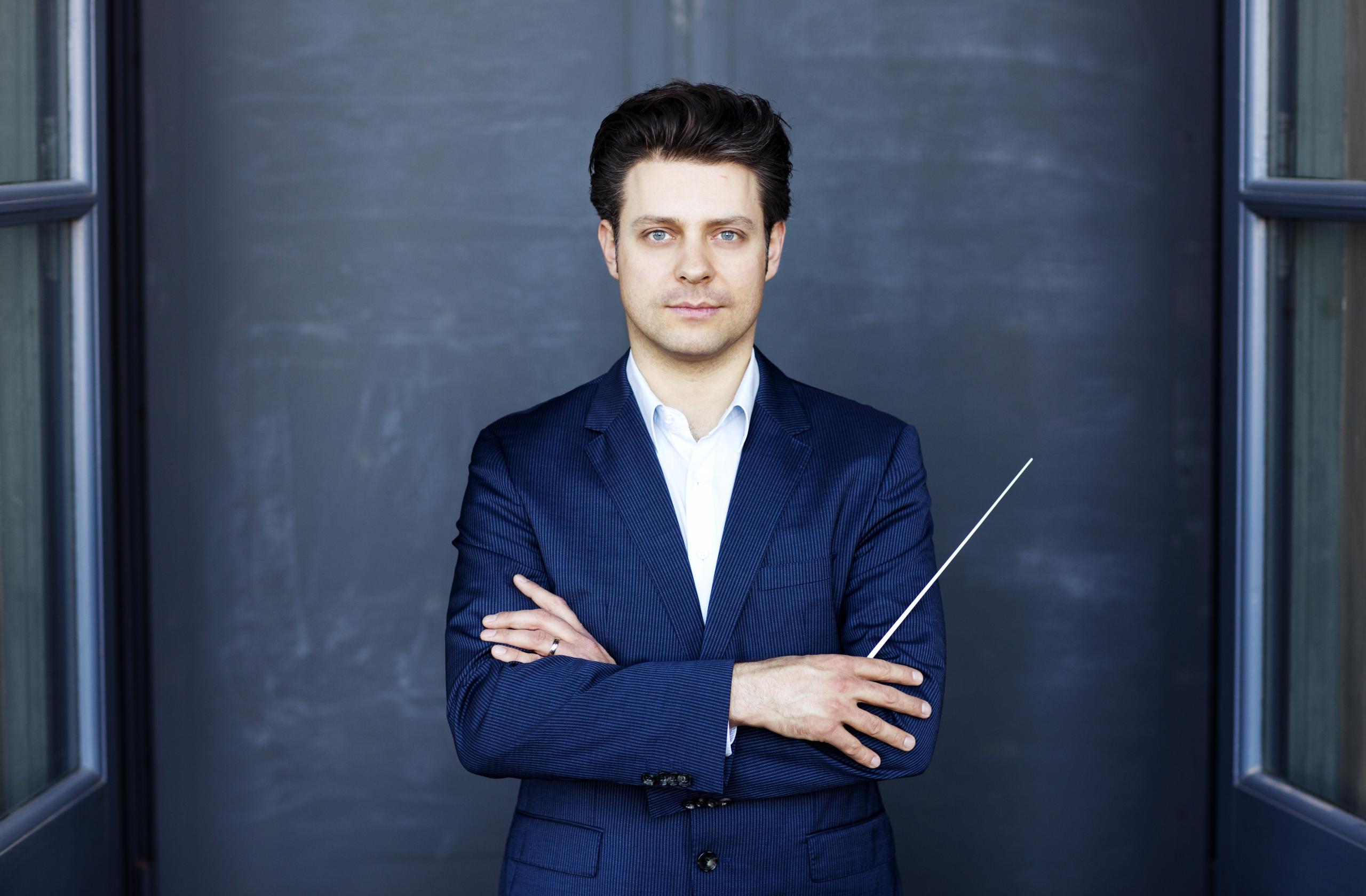 Portrait of the conductor Joseph Bastian. He is wearing a dark blue jacket and is depicted against a grey background.