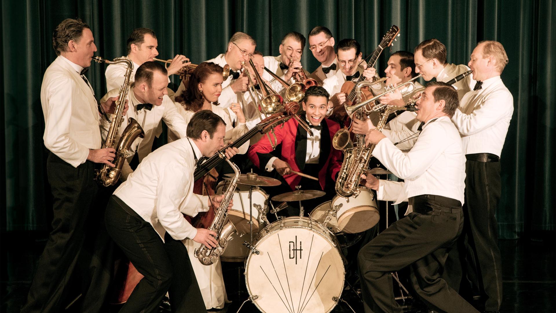Man sieht die Mitglieder des Swing Dance Orchestras, wie sie mit ihren Instrumenten rund um einen Schlagzeuger stehen. Alle tragen Anzüge im Stil der 20er-Jahre. 