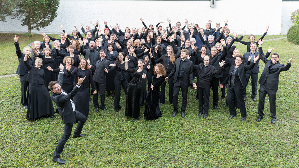 The members of the choir on a green field, all raise their hands and wave.