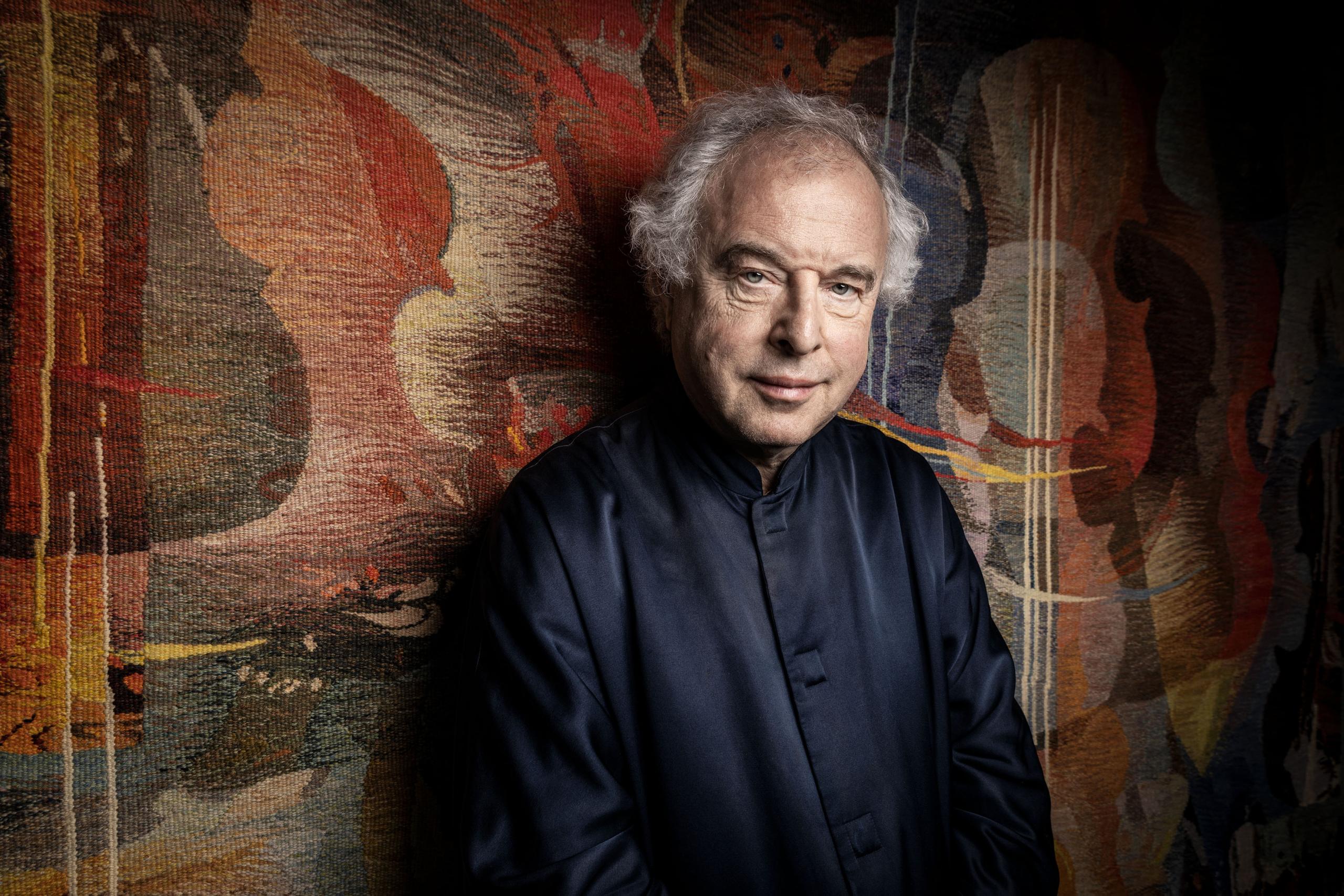 Portrait of the pianist and conductor Sir András Schiff. He wears a black shirt and stands in front of a patterned wall.