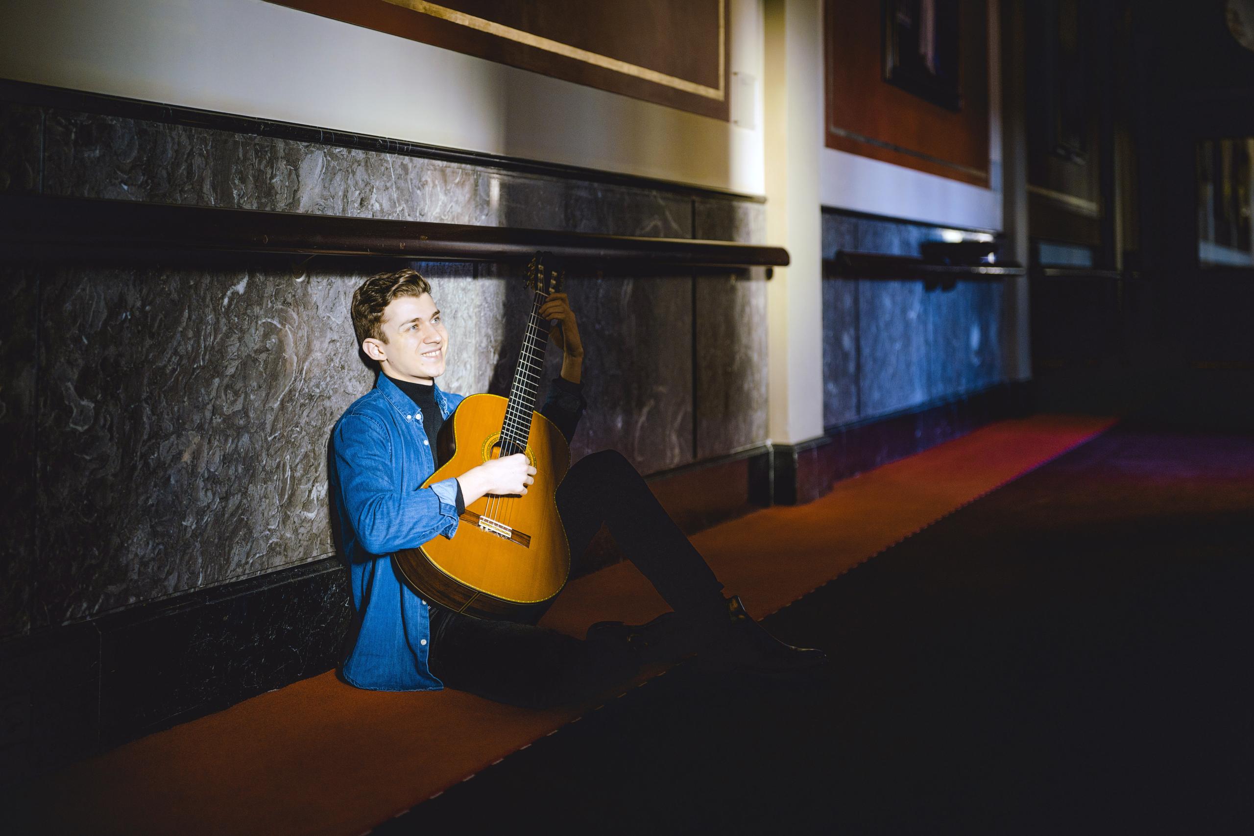 Portrait of the guitarist Thibaut Garcia. He is sitting with his guitar on the floor of a shady corridor.