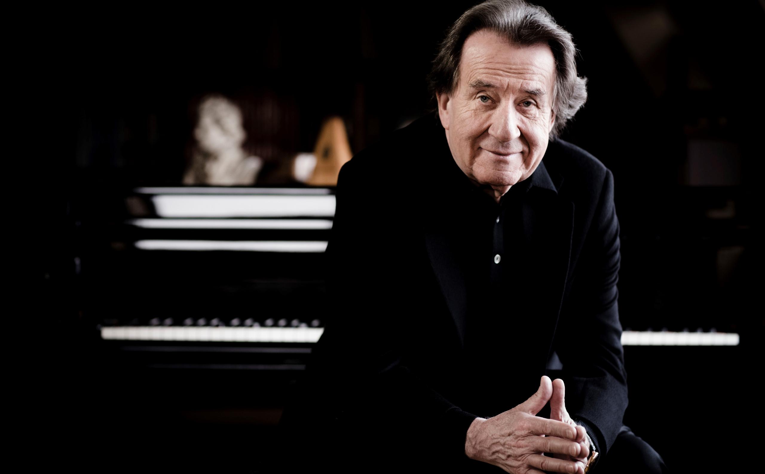 Portrait of the pianist Rudolf Buchbinder. He sits in front of a piano in a black suit and looks into the camera.