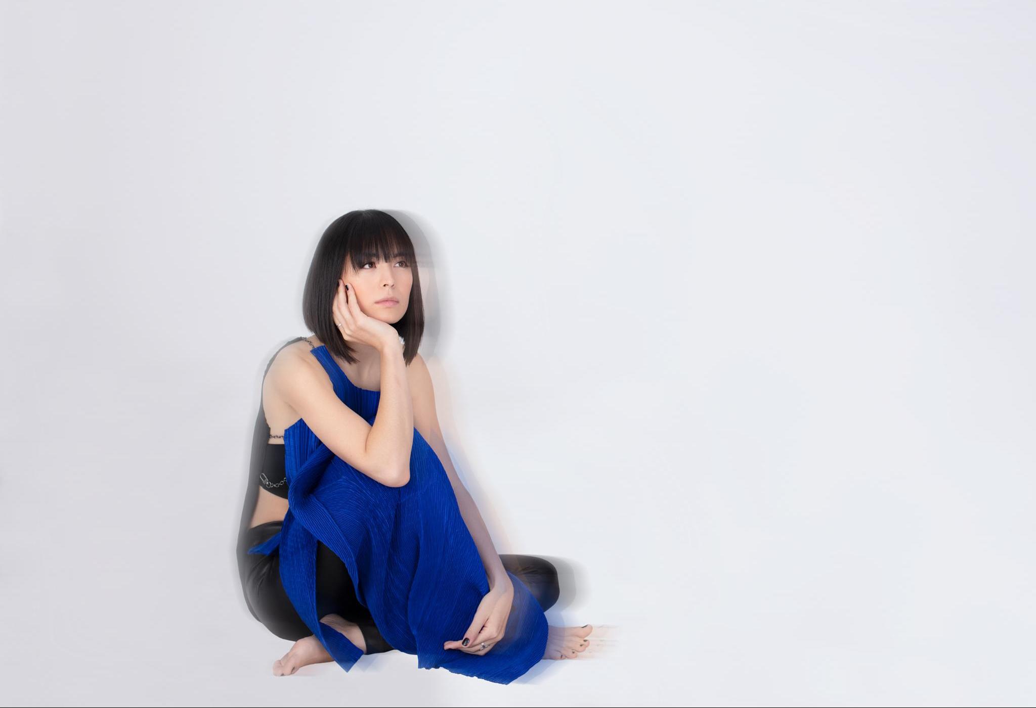 Porträt der Pianistin Alice Sara Ott. Sie sitzt in einem blauen Kleid vor einem weißen Hintergrund. Das Bild ist so bearbeitet, dass es leicht verschwommen wirkt.