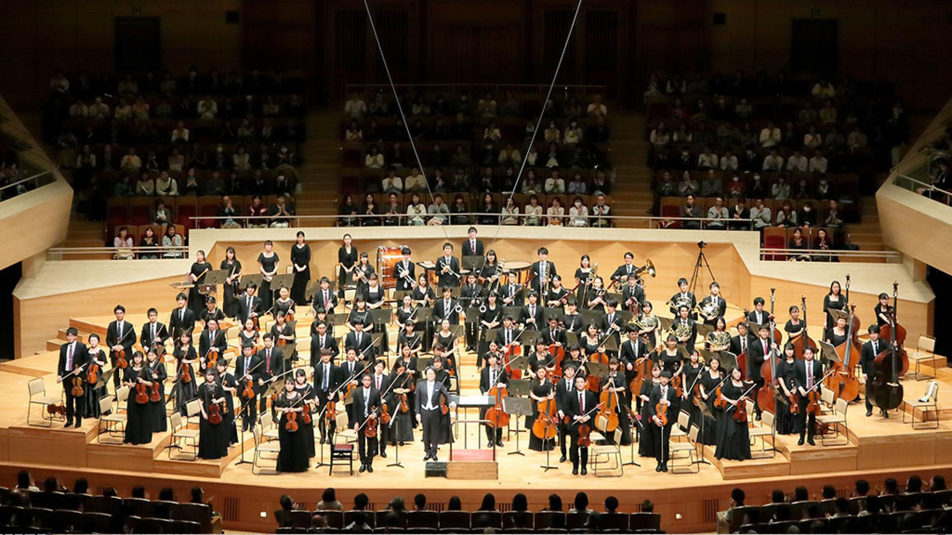 The Waseda Symphony Orchestra Tokyo is depicted on a stage. The musicians are dressed in black and look into the auditorium while standing.