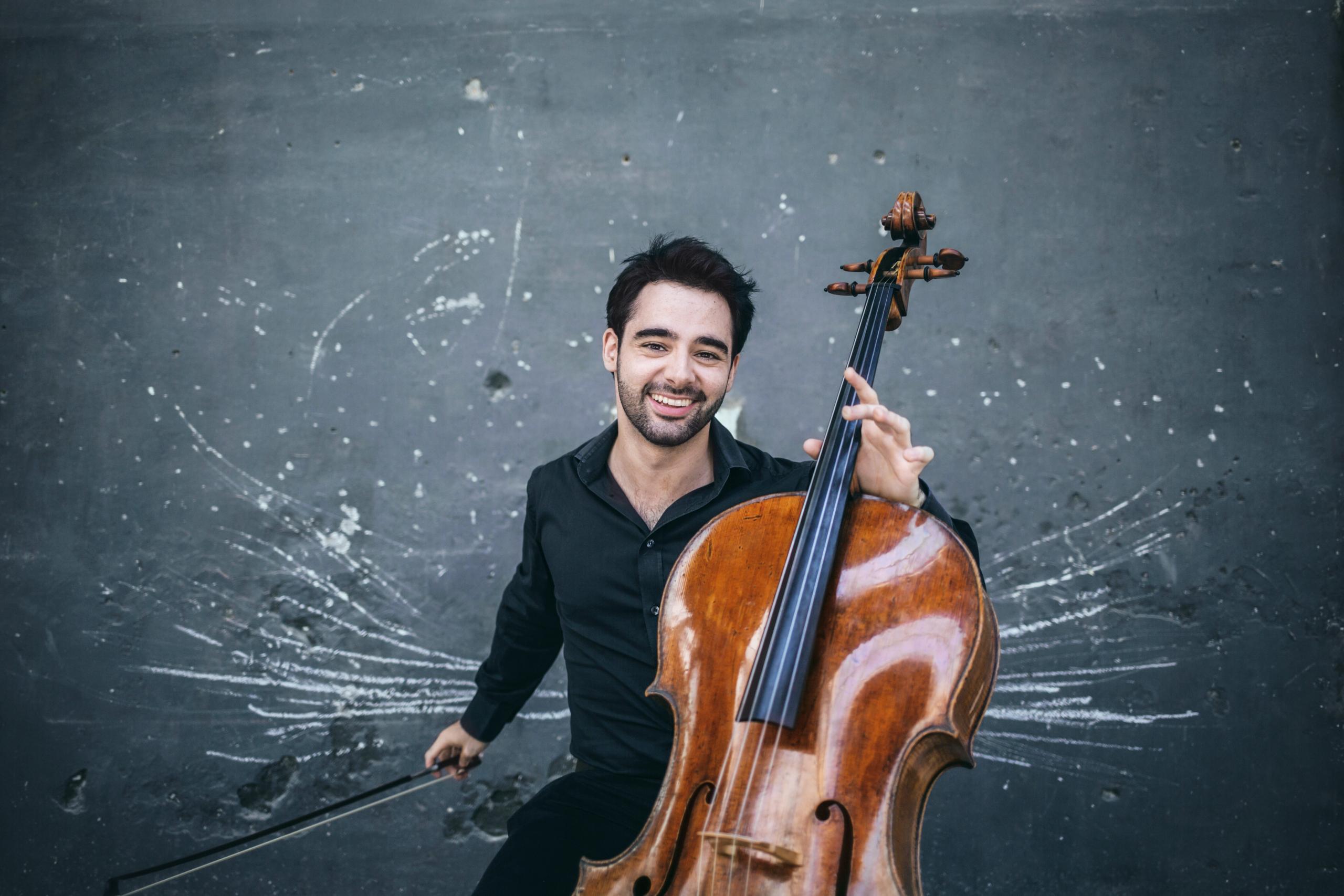 Porträt des Cellisten Pablo Ferrández. Er hält sein Cello in der Hand und sitzt vor einer grauen Wand, die mit weißer Kreide bemalt ist.