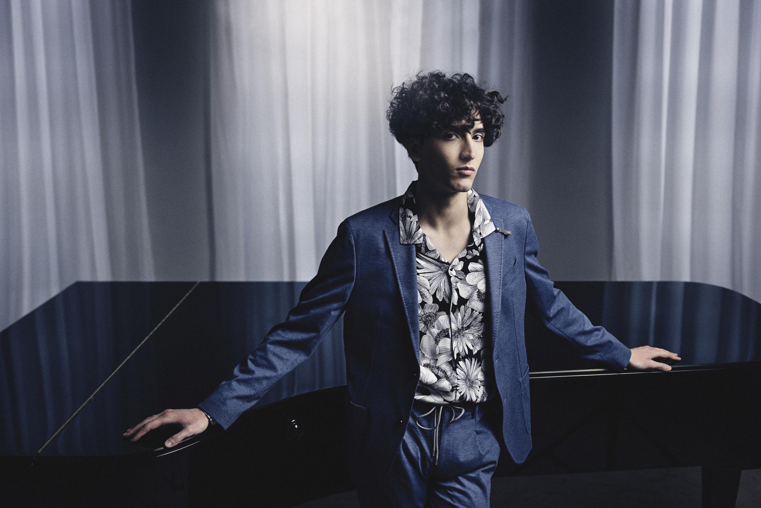 Portrait of the pianist Roman Borisov. He is standing in front of a grand piano in a blue suit with a patterned shirt.