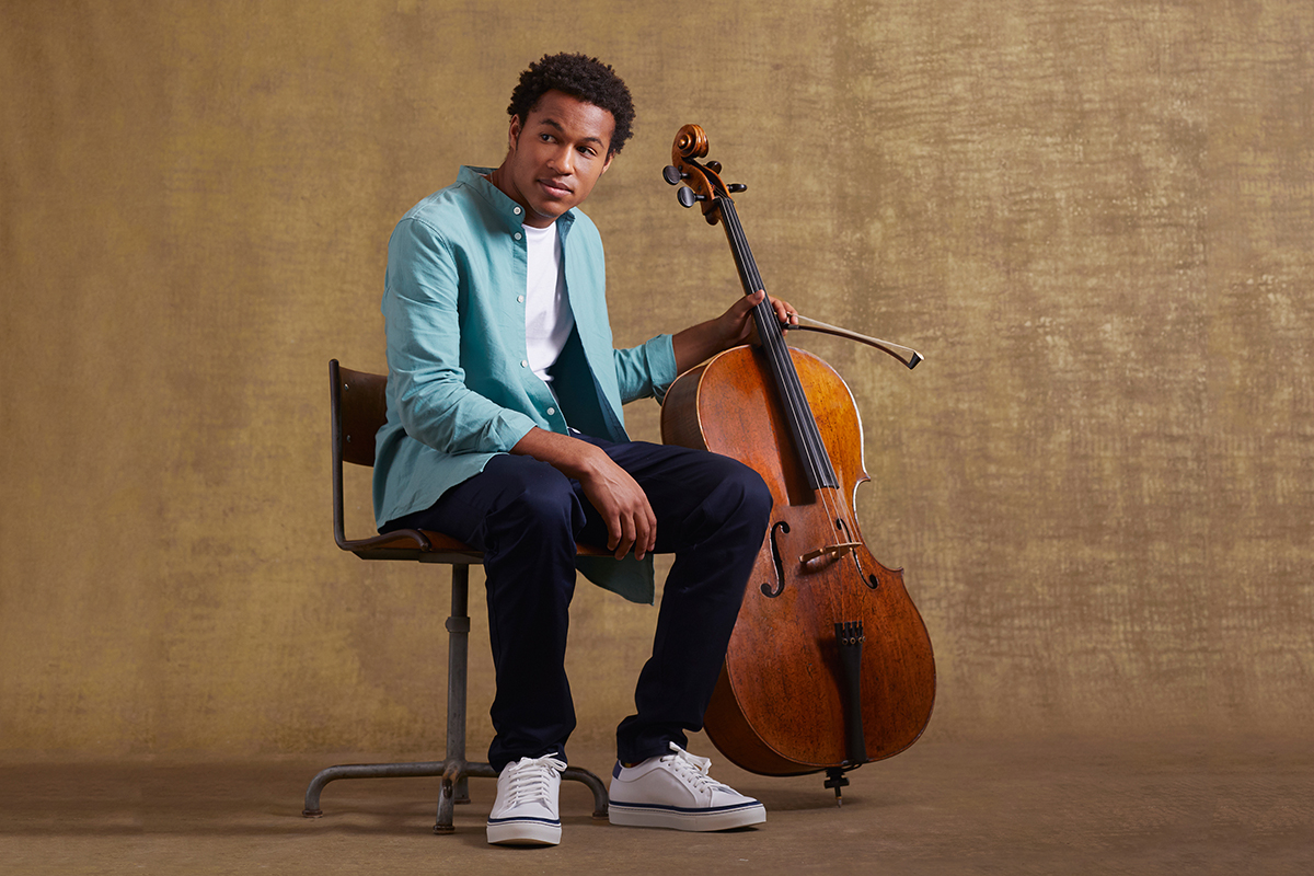 Portrait of the cellist Sheku Kanneh-Mason. He wears a casual outfit, holds his cello in his hand and sits in front of a brown background.