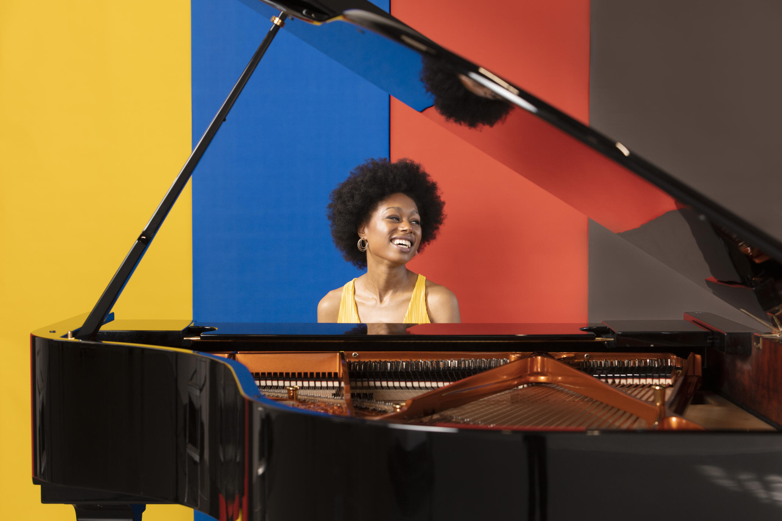 Portrait of the pianist Isata Kanneh-Mason. She is sitting behind a grand piano in a yellow dress, laughing. The wall behind her is striped in yellow, blue, red and brown.