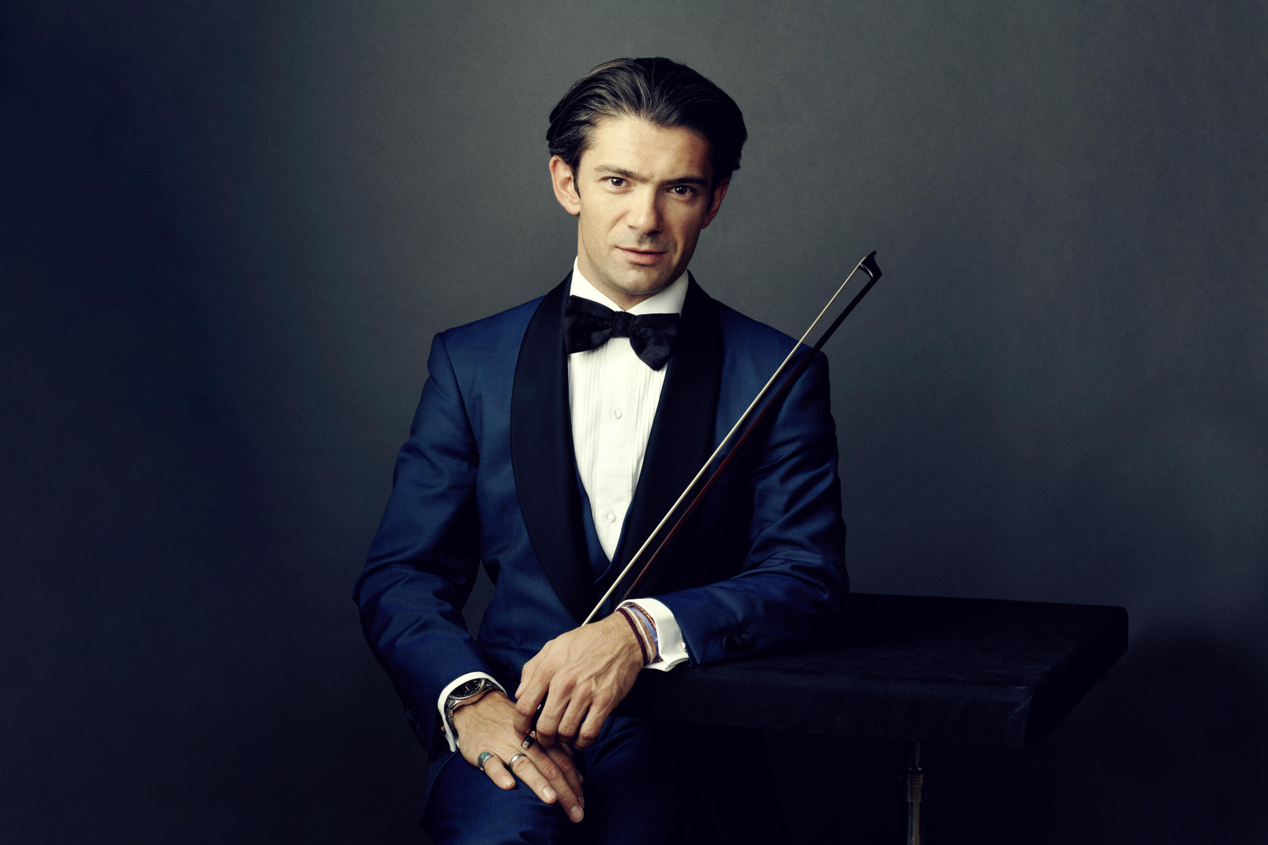 Portrait of the cellist Gautier Capuçon. He is wearing a blue jacket and is sitting in front of a grey wall. 