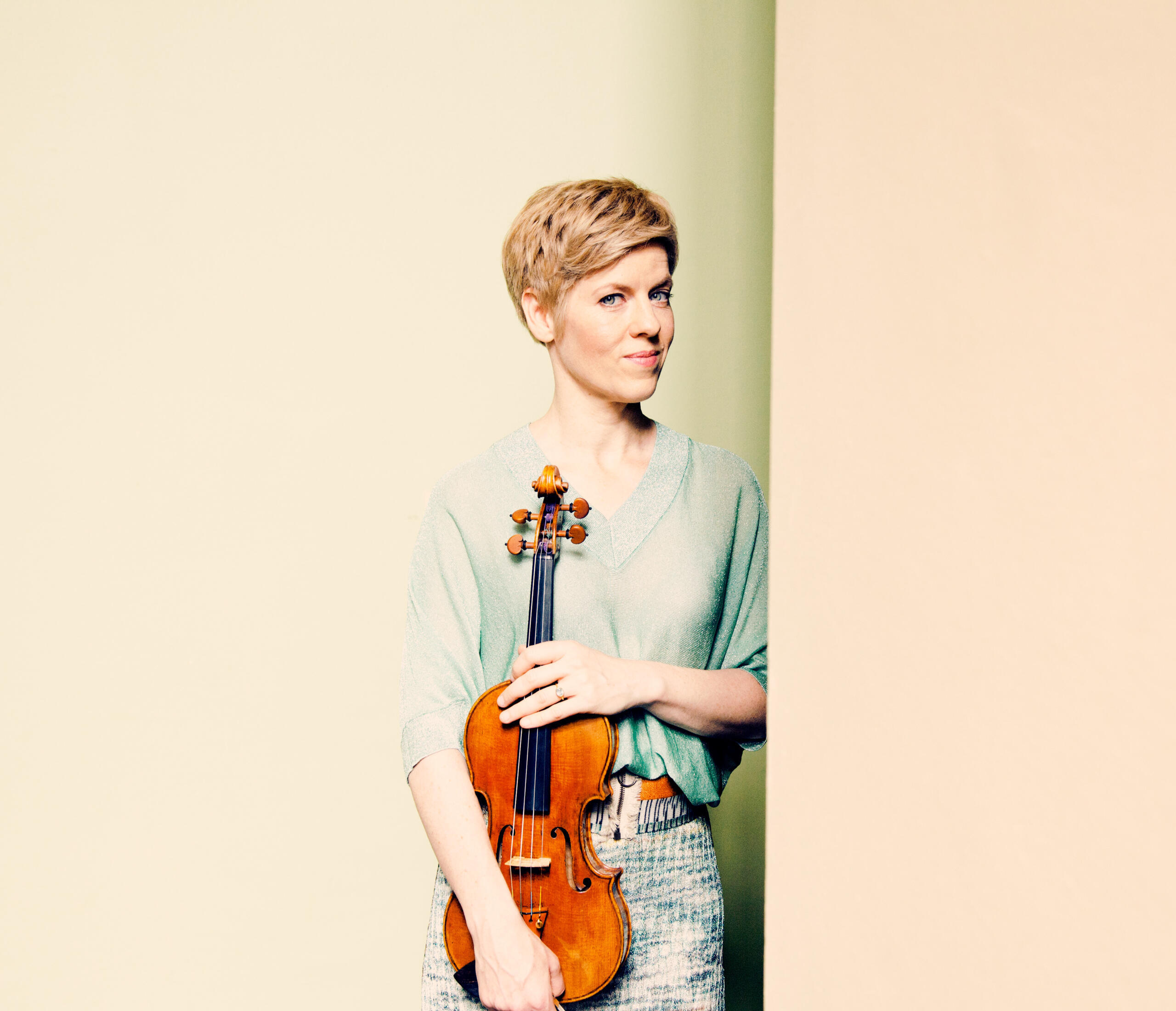 Portrait of the violinist Isabelle Faust. She holds her violin in her hand and stands leaning against a beige wall.