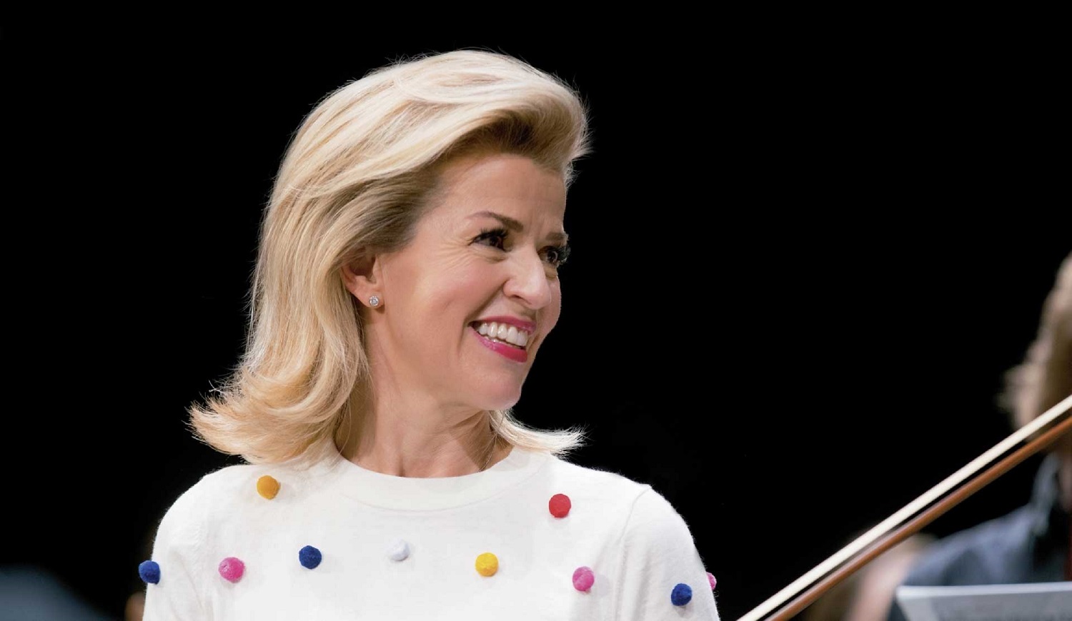 Anne-Sophie Mutter can be seen in side profile. She is wearing a white top with coloured dots and is laughing.