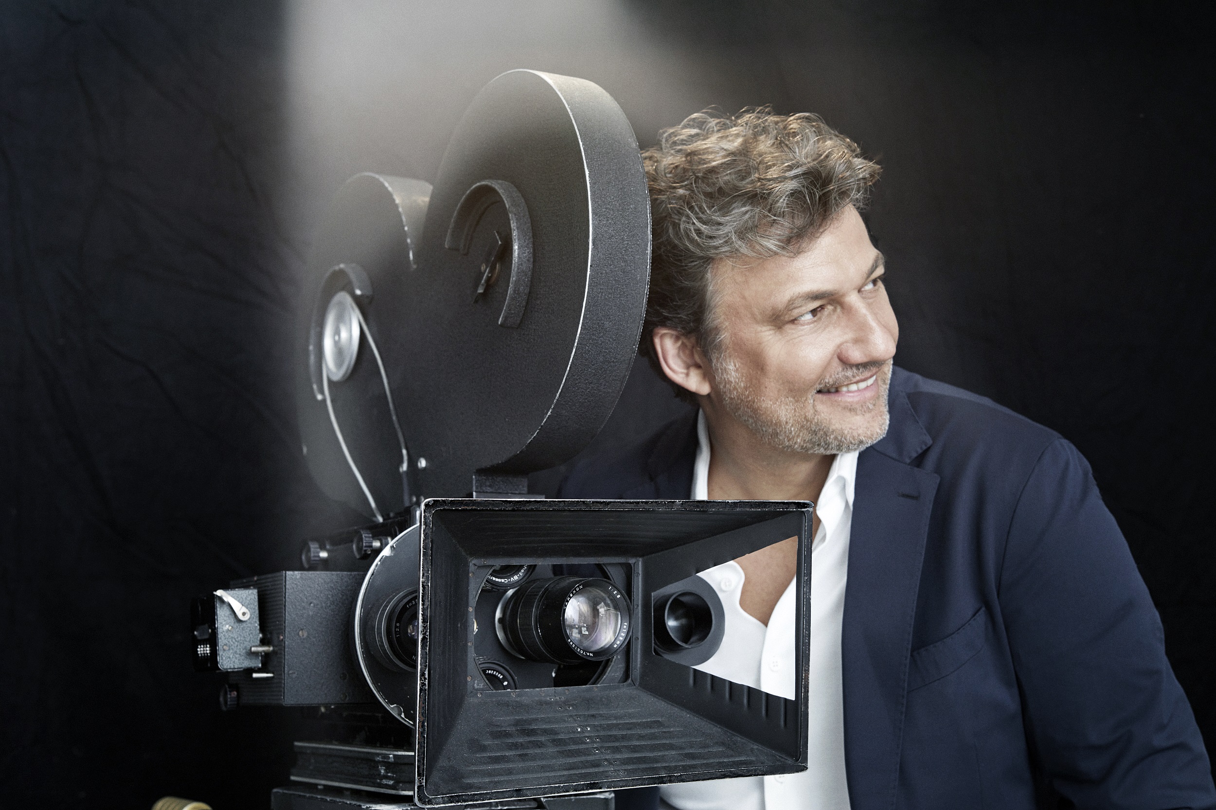 Jonas Kaufmann is pictured next to an old film projector. He is seen in side profile, smiling and wearing a white shirt and a dark blue jacket.