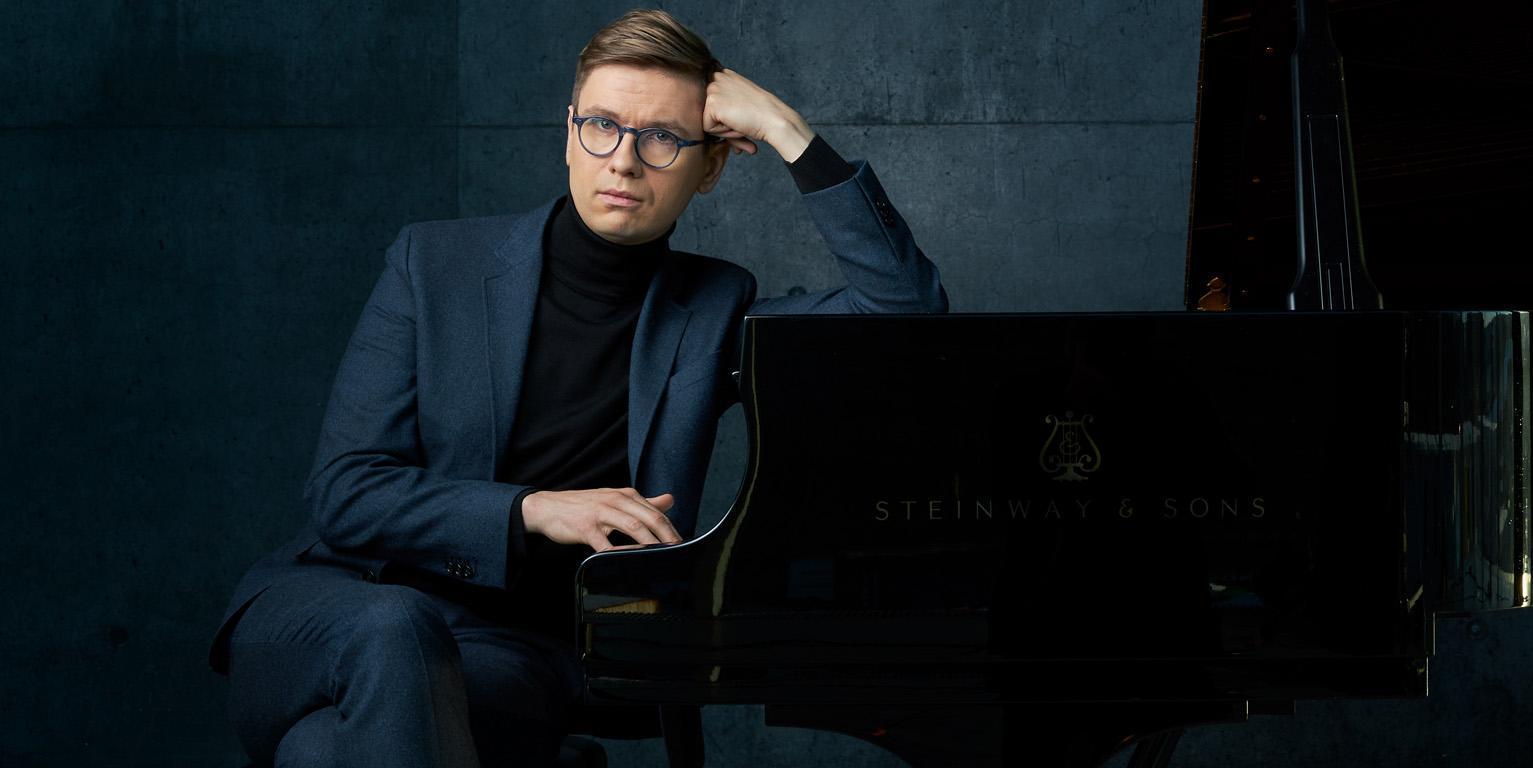 Víkingur Ólafsson sits in front of a grand piano, resting his elbow on it
