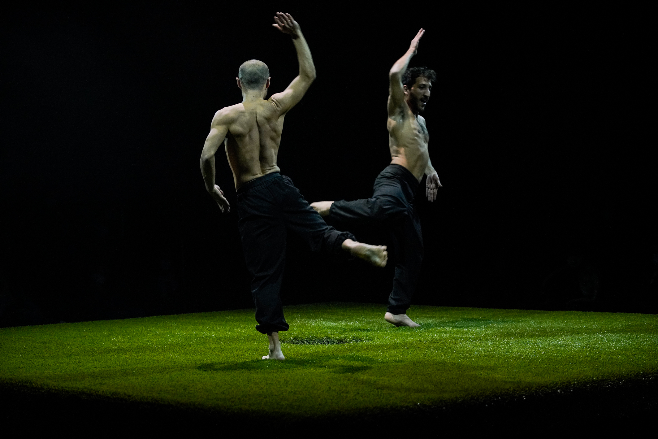 Zwei Tänzer stehen mit schwarzen Hosen und ohne Oberteil tanzend auf einer Grasfläche, die auf einer Bühne ausgebreitet ist.