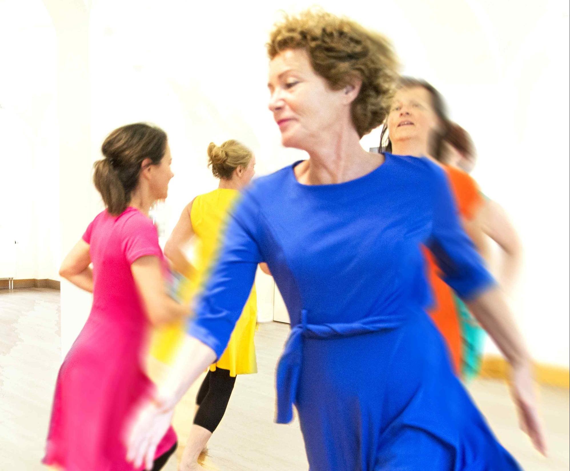 Several women in brightly colored dresses walk in a circle, in the foreground is a lady in a royal blue dress