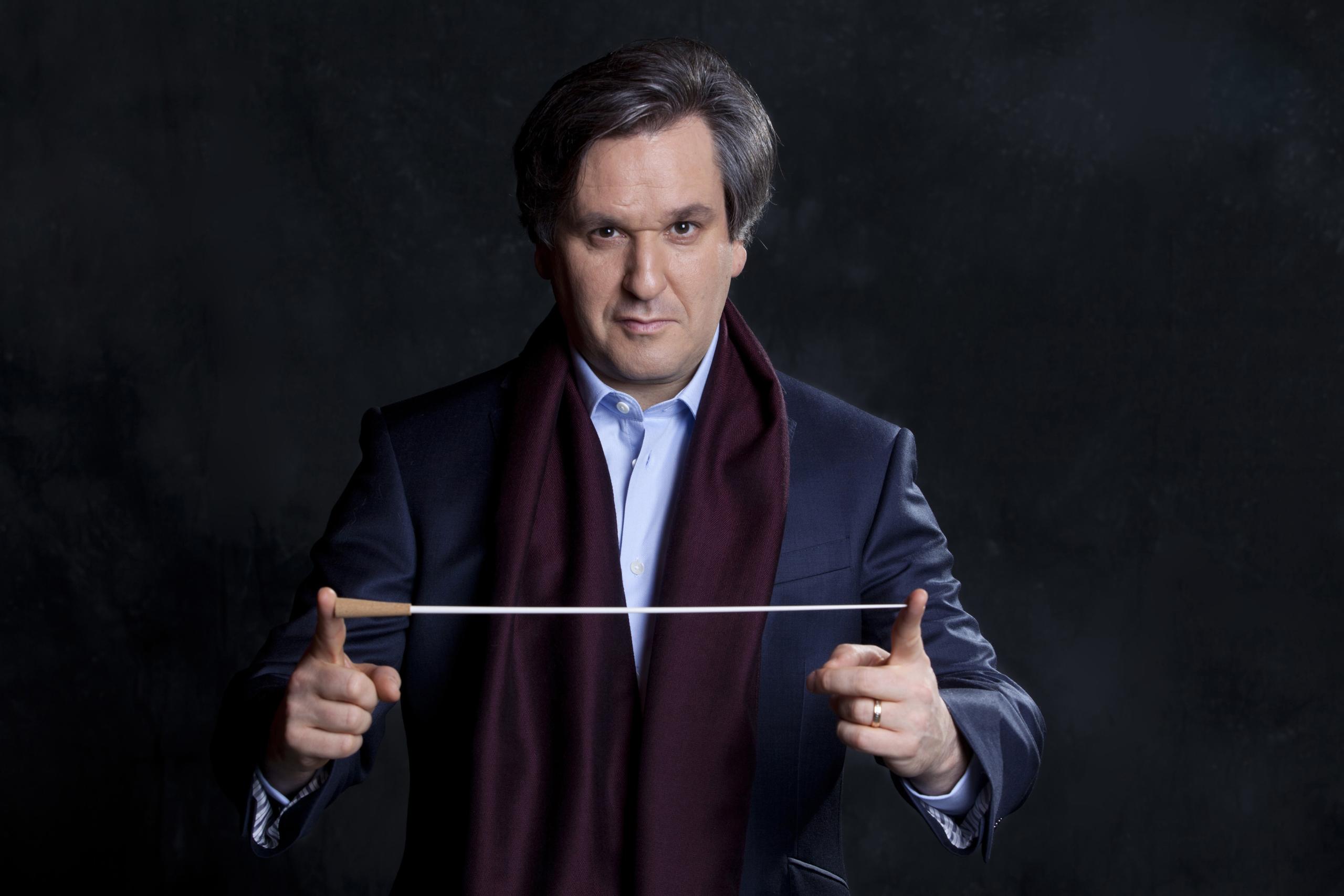 Der Dirigent präsentiert seinen Stab. Er steht vor einem dunklen Hintergrund, trägt einen blauen Anzug und einen weinroten Schal.