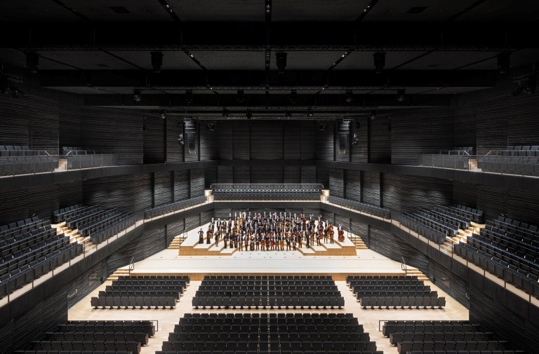 Leerer Konzertsaal mit schwarzen Wänden und Sitzen, hellem Boden, auf der Bühne stehend die Musiker und Musikerinnen der Münchner Philharmoniker