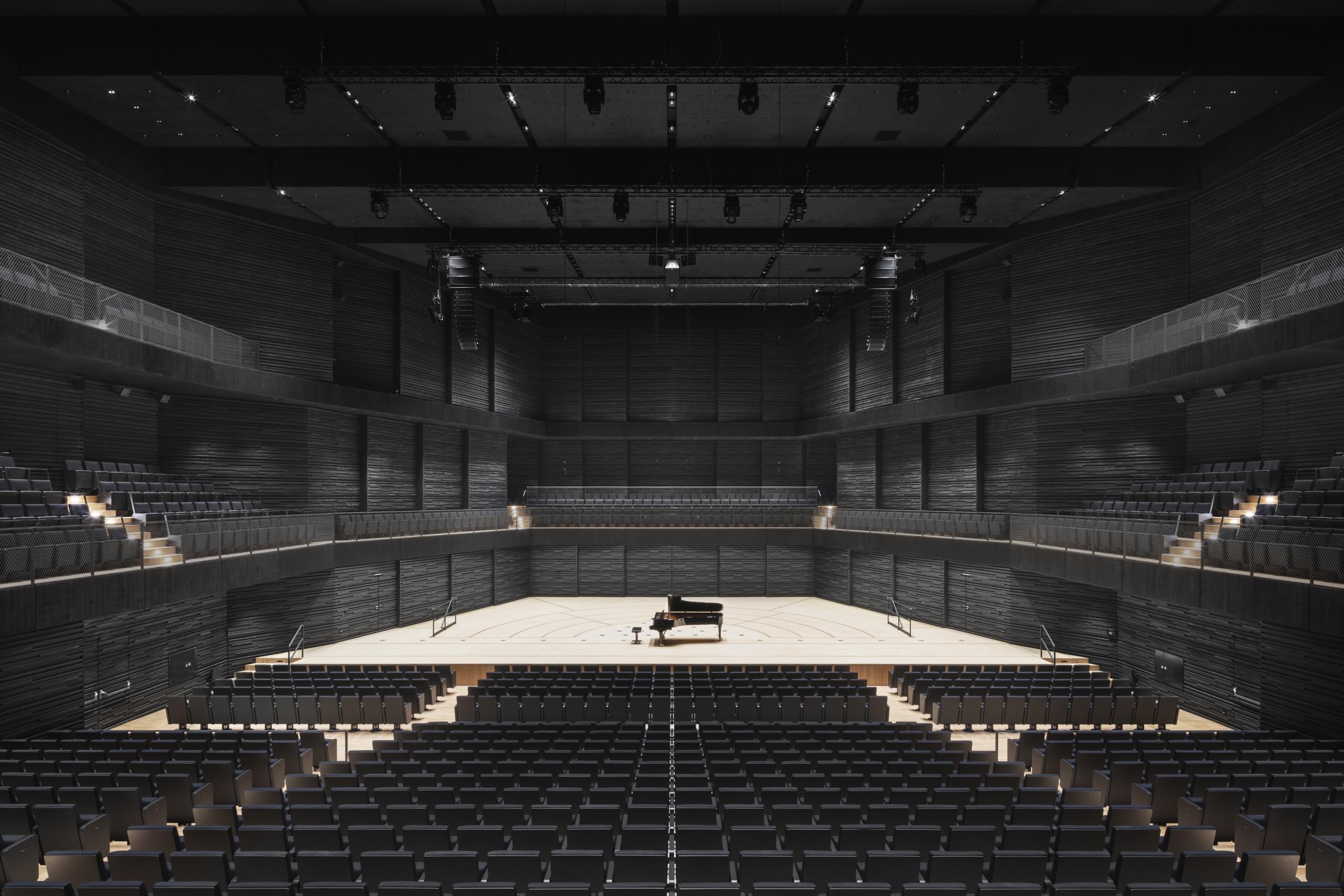 Empty concert hall with black wood paneling, the floor and stage are light wood, black seating, on the stage a grand piano