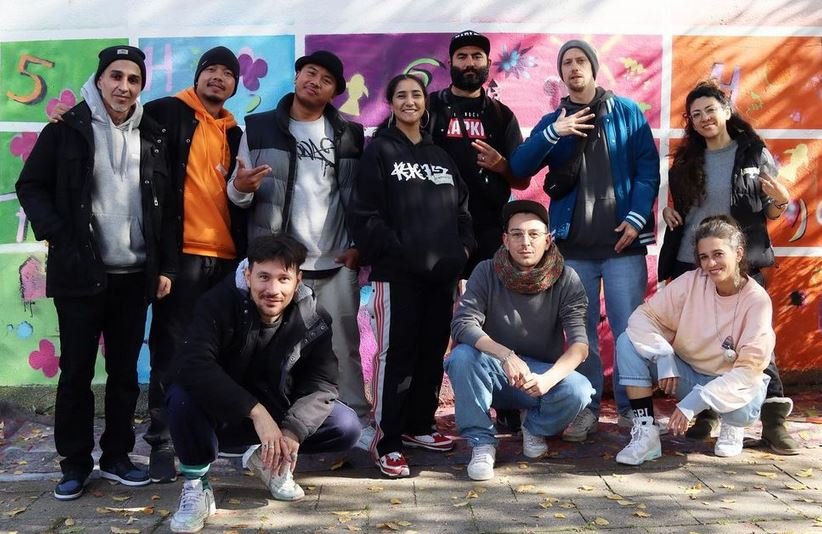 Eine Gruppe junger Menschen im Hip-Hop-Stil bekleidet vor einer Graffitti-Wand. 