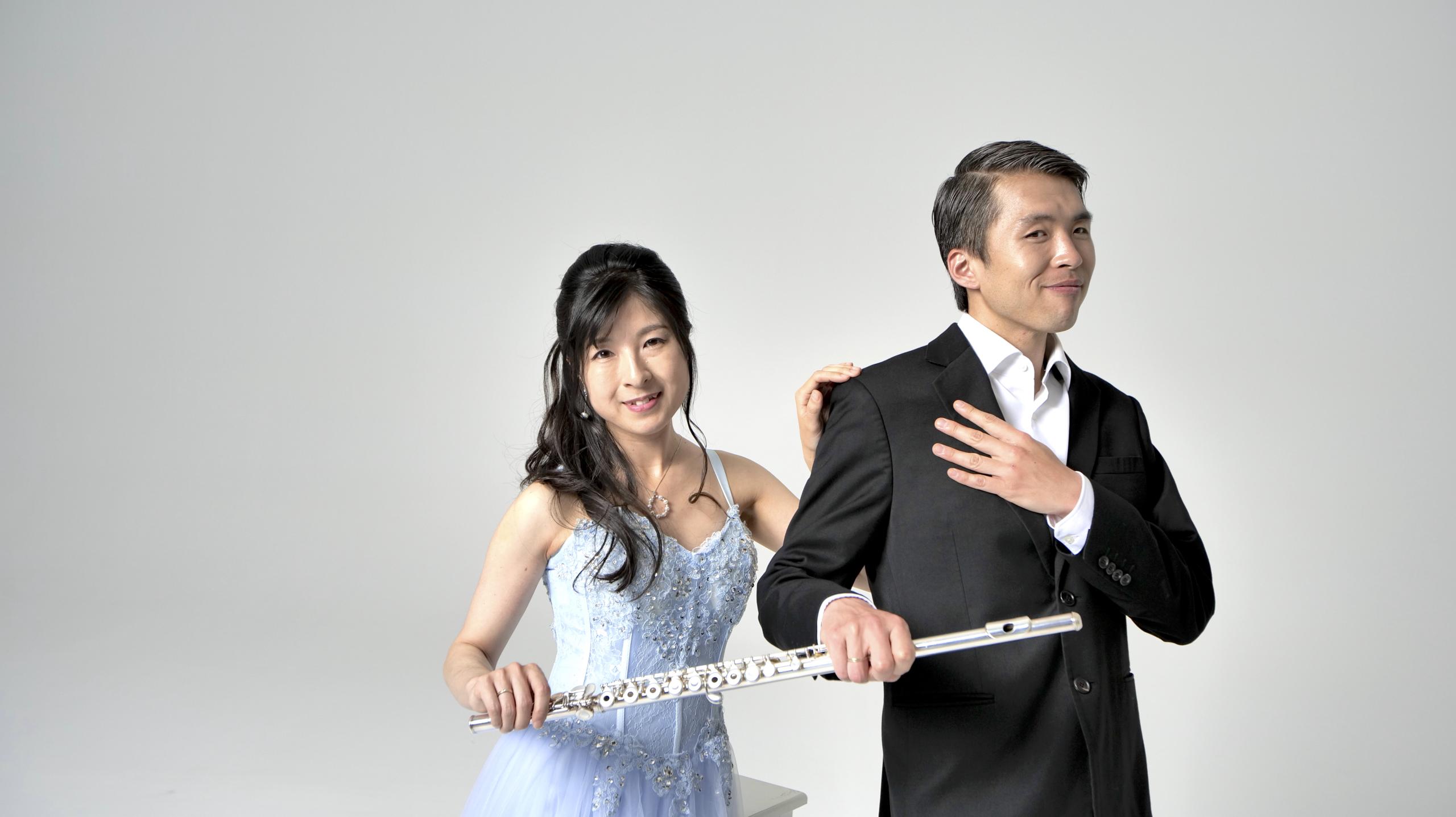 A young woman in a light blue evening dress, she puts her hand on the shoulder of a young man in a tailcoat. Both hold a flute to the front.
