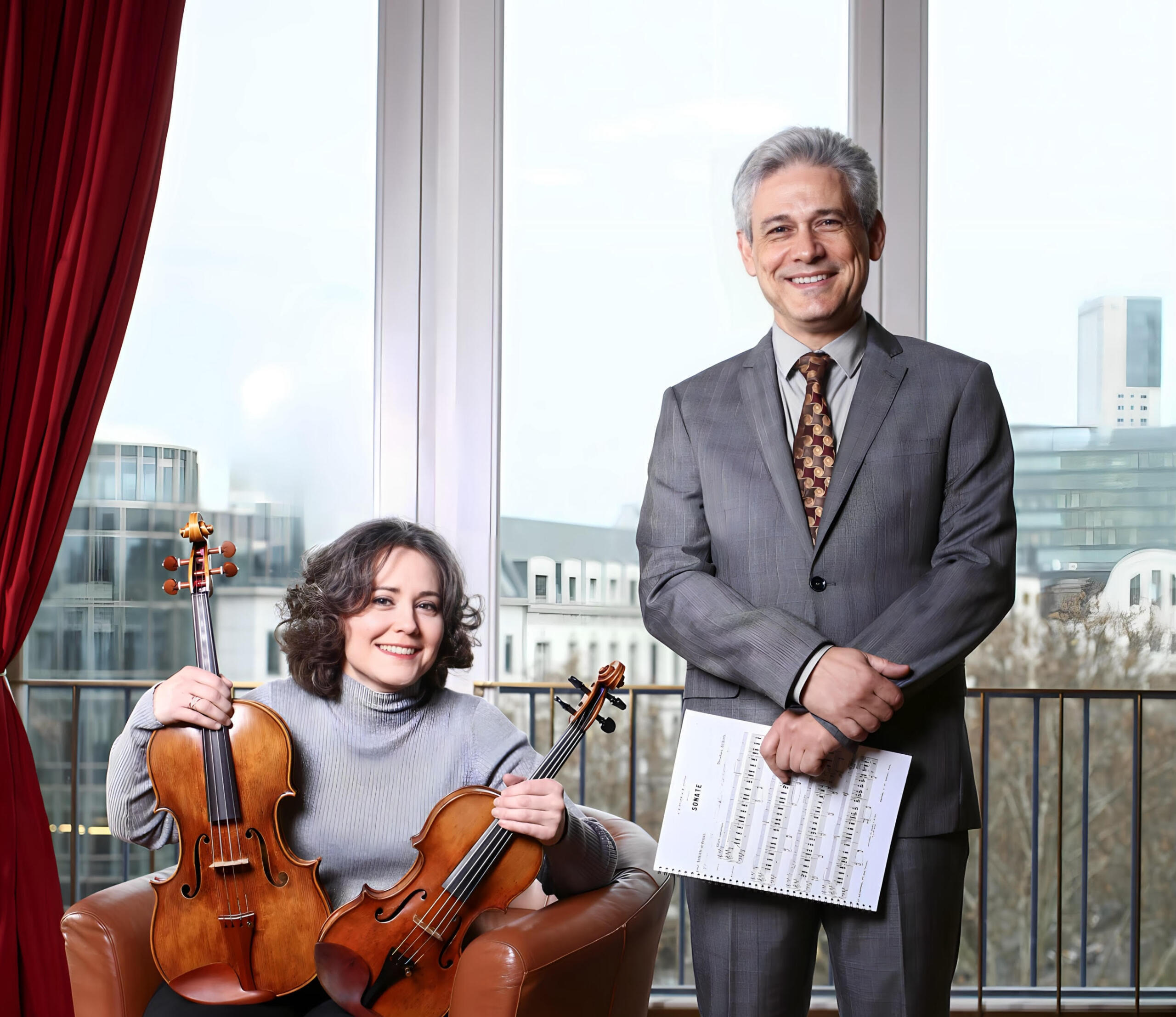 A woman sits on a leather armchair holding a violin and a viola. Next to her is a man in a grey suit. Both are smiling at the camera.
