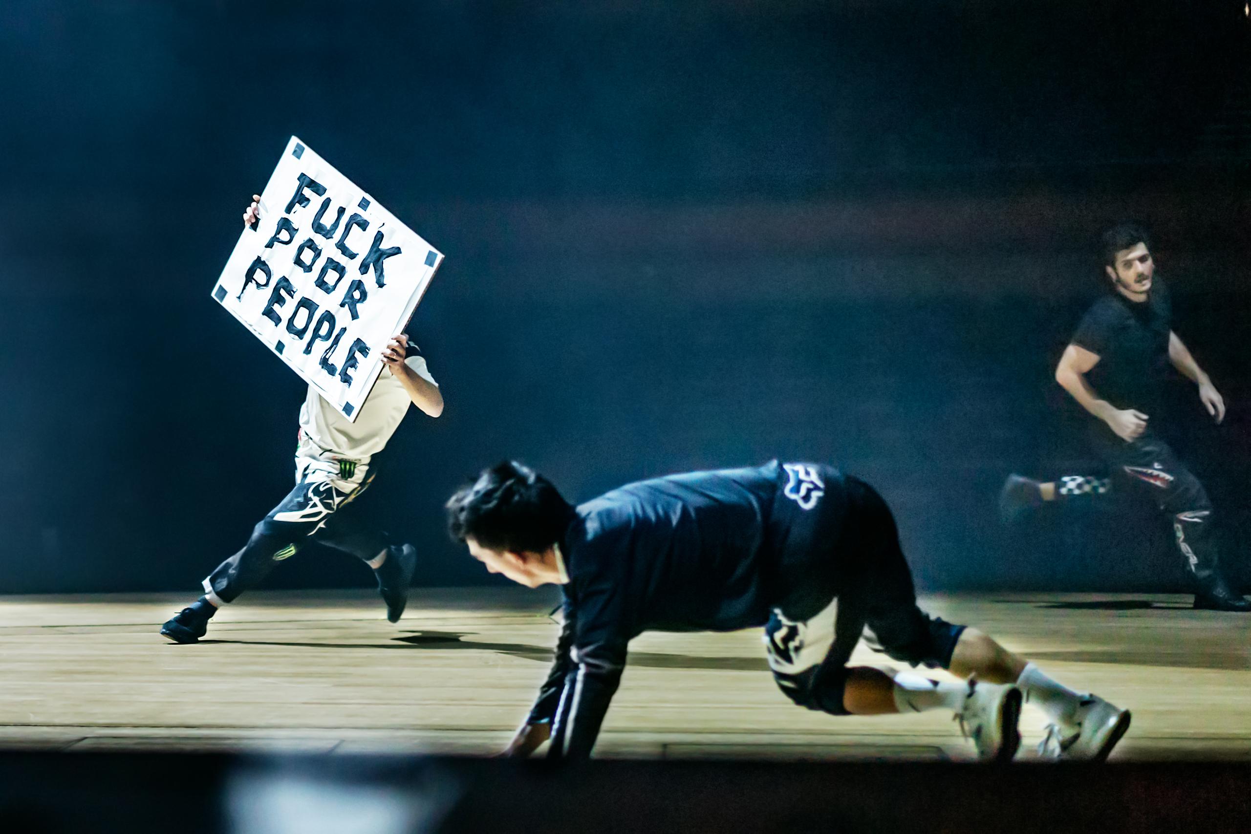 A stage with a dark background. On it three men running. One of them carries a sign with the inscription "Fuck Poor People".