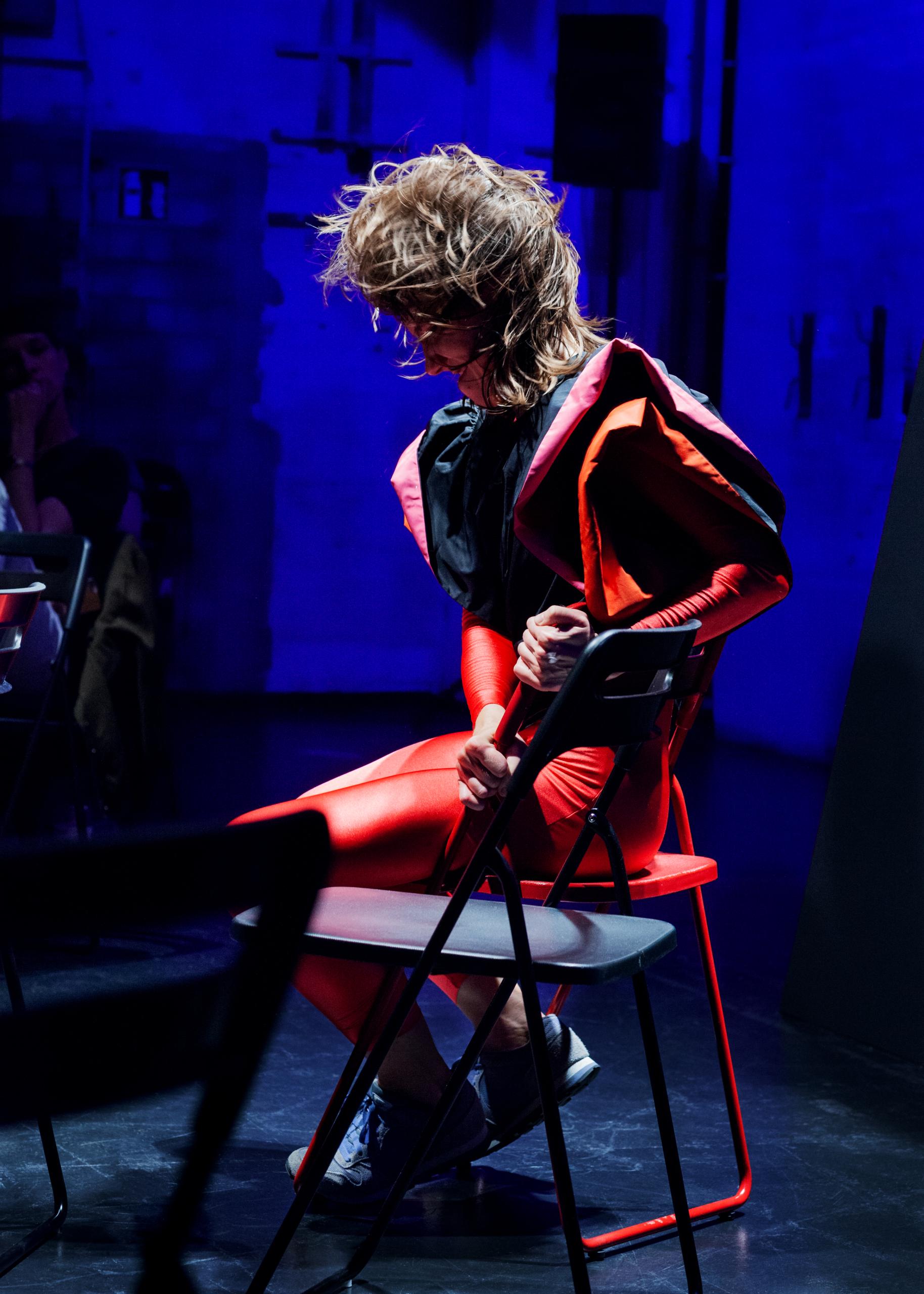 Blue lit stage, woman in red costume sits on folding chair