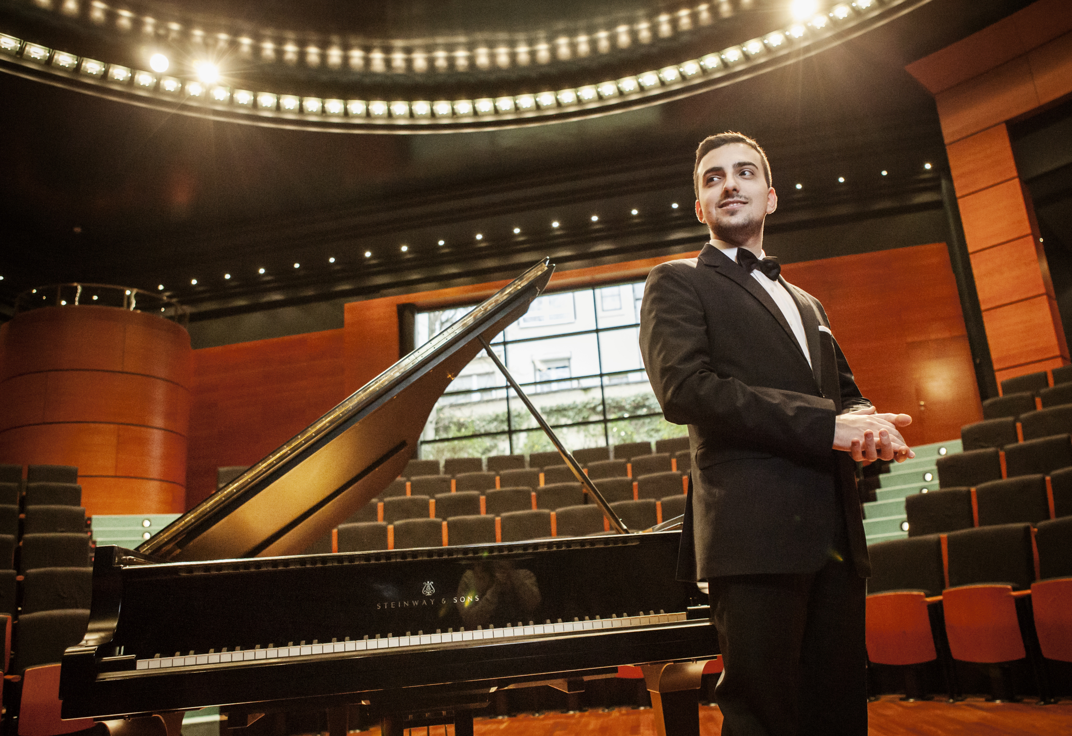 A small concert hall with empty seats. The pianist Ferro stands in front of a grand piano in the middle of the room.