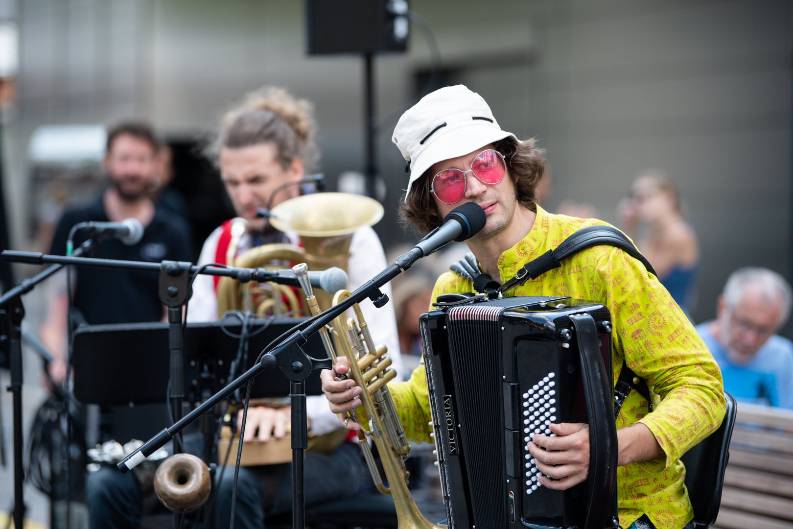 Zwei Musiker mit Mikros, mehreren Instrumenten: Akkordeon, Trompete, Tuba. Ein Musiker trägt einen Hut und eine rosarote Sonnenbrille.