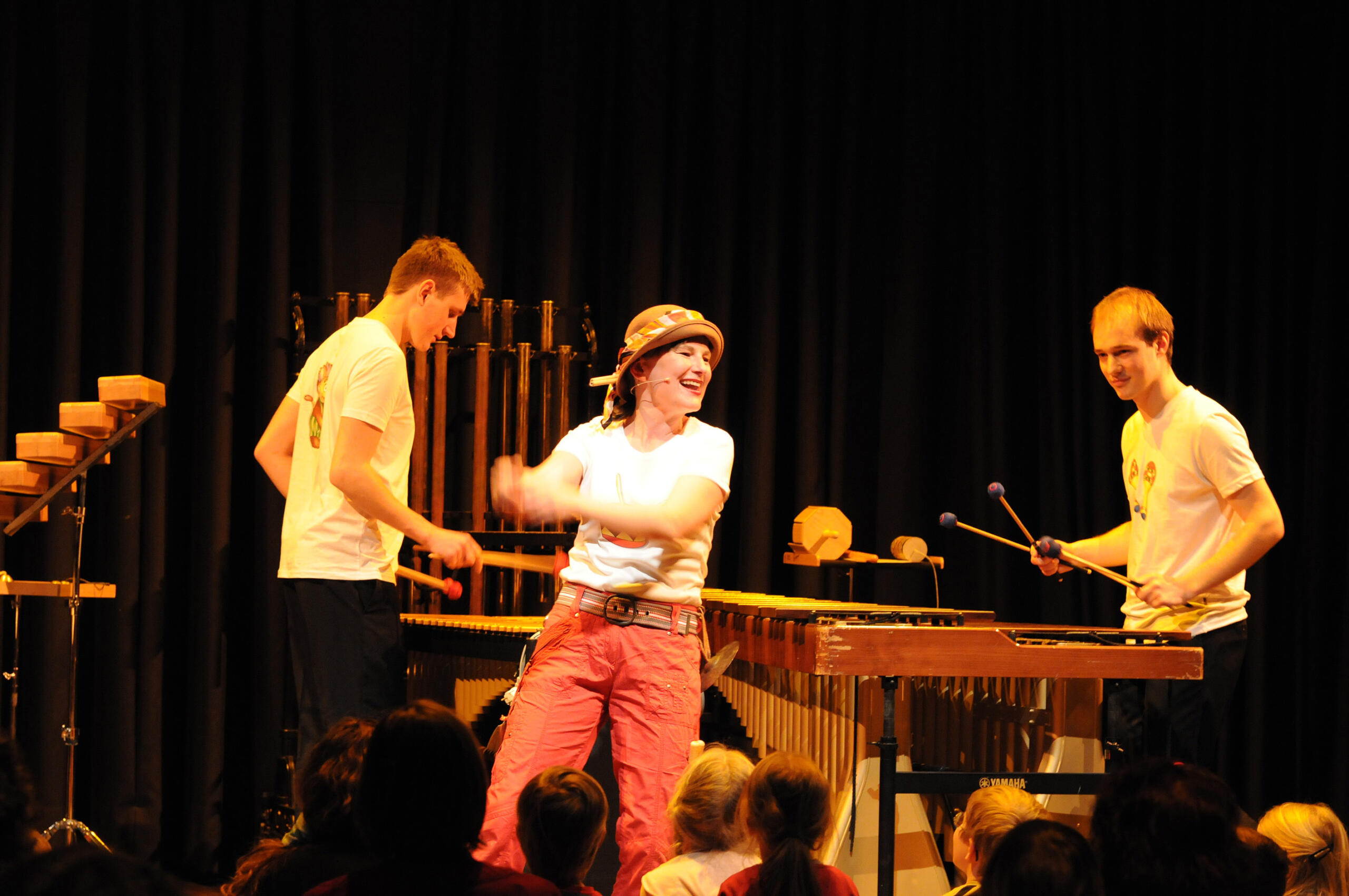 Eine Frau steht auf einer Bühne und tanzt und lacht. Im Hintergrund sieht man zwei Schlagwerker an ihren Instrumenten.