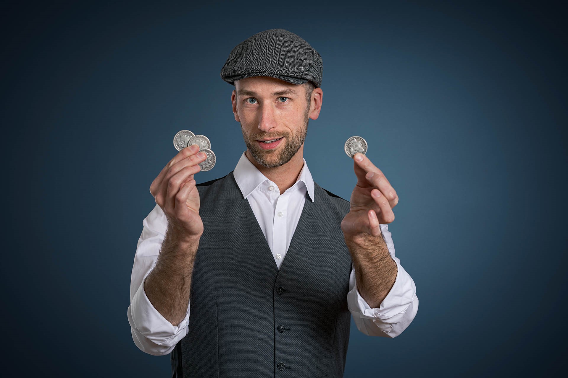 Ein Mann in Anzug und mit einer Baskenmütze steht vor einem grauen Hintergrund. In der Hand hält er silberne Münzen.
