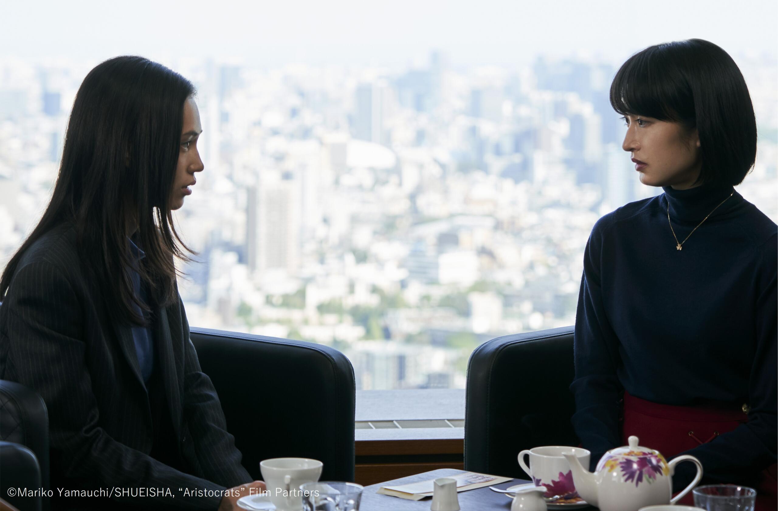 Zwei Frauen in schwarzer Kleidung sitzen sich gegenüber. Vor ihnen steht eine Teeservice. Hinter ihnen sieht man ein Fenster und dahinter die Skyline einer großen Stadt.