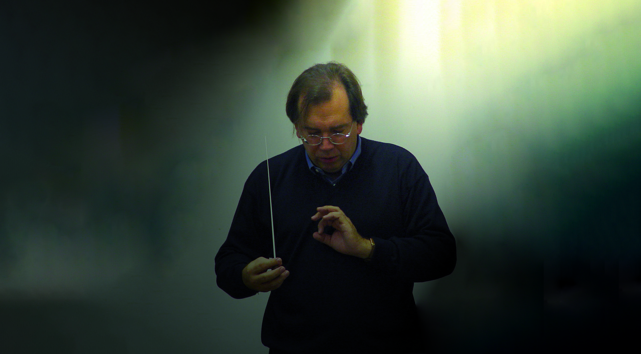 Porträt des Dirigenten Christian Kabitz. Er steht in einem lichtdurchfluteten Raum, hält einen Dirigentenstab in der Hand und ist ganz in sein Dirigat vertieft.