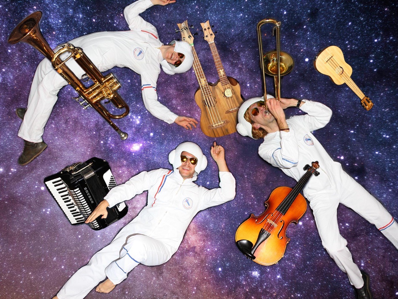 Drei Musikerinnen liegen in weißen Astronauten-Anzügen im lilafarbenen Kosmos, umgeben von verschiedenen Instrumenten.