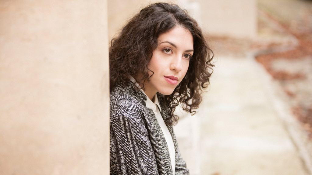 Portrait shot of the pianist Beatrice Rana. She has brown curls and leans against a beige house wall.