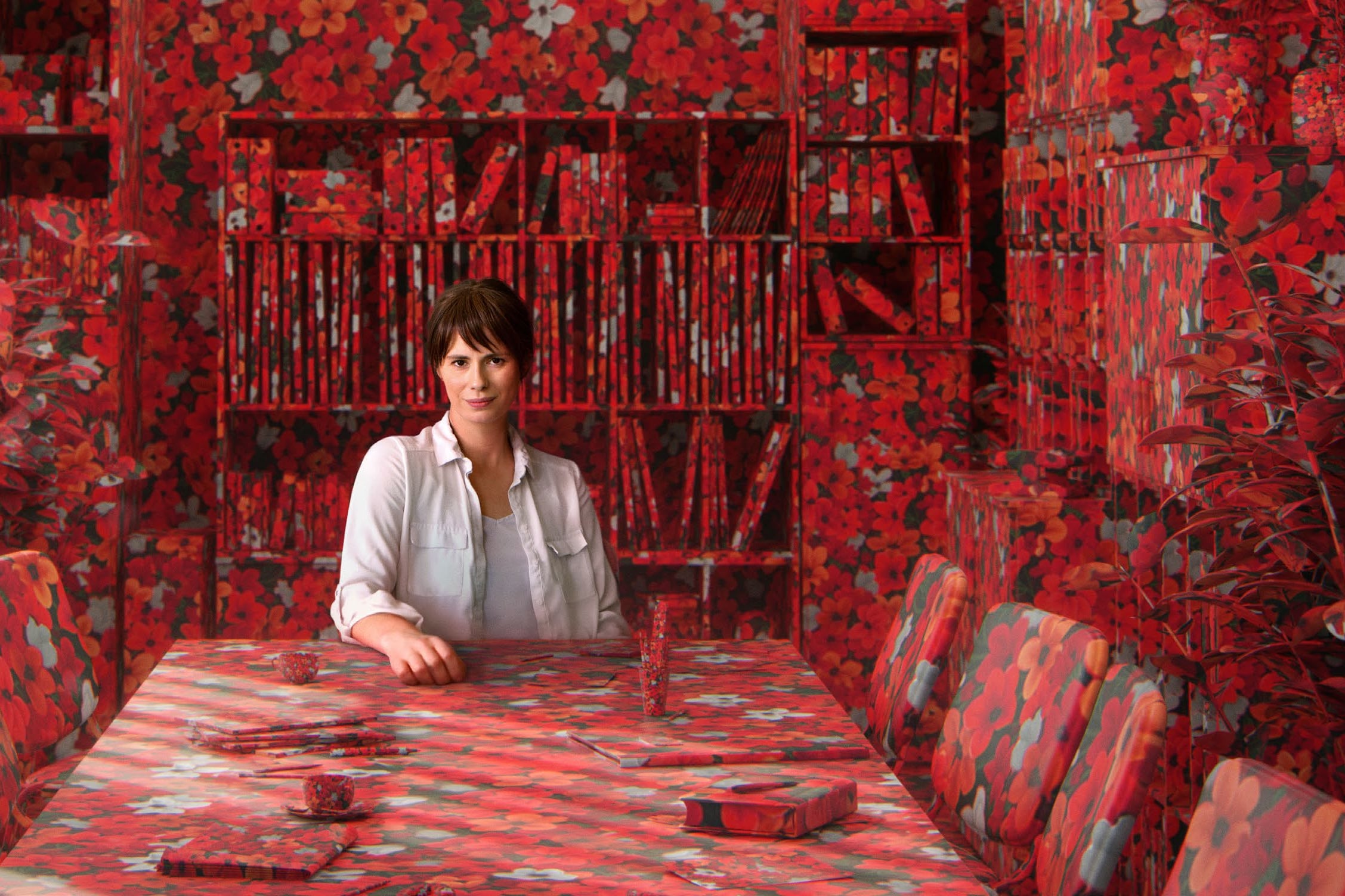 Eine Frau sitzt an einem Tisch, der mit einer Tischdecke mit roten Rosen bedeckt ist. Dasselbe Muster findet sich auf den Stühlen und an den Wänden des Raumes wieder.