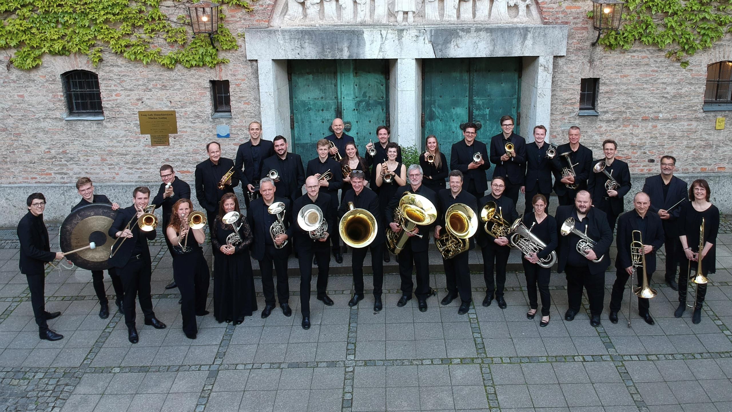 Foto der der Mitglieder der Brass Band München mit ihren Instrumenten vor der Senndlinger Himmelfahrtkirche.