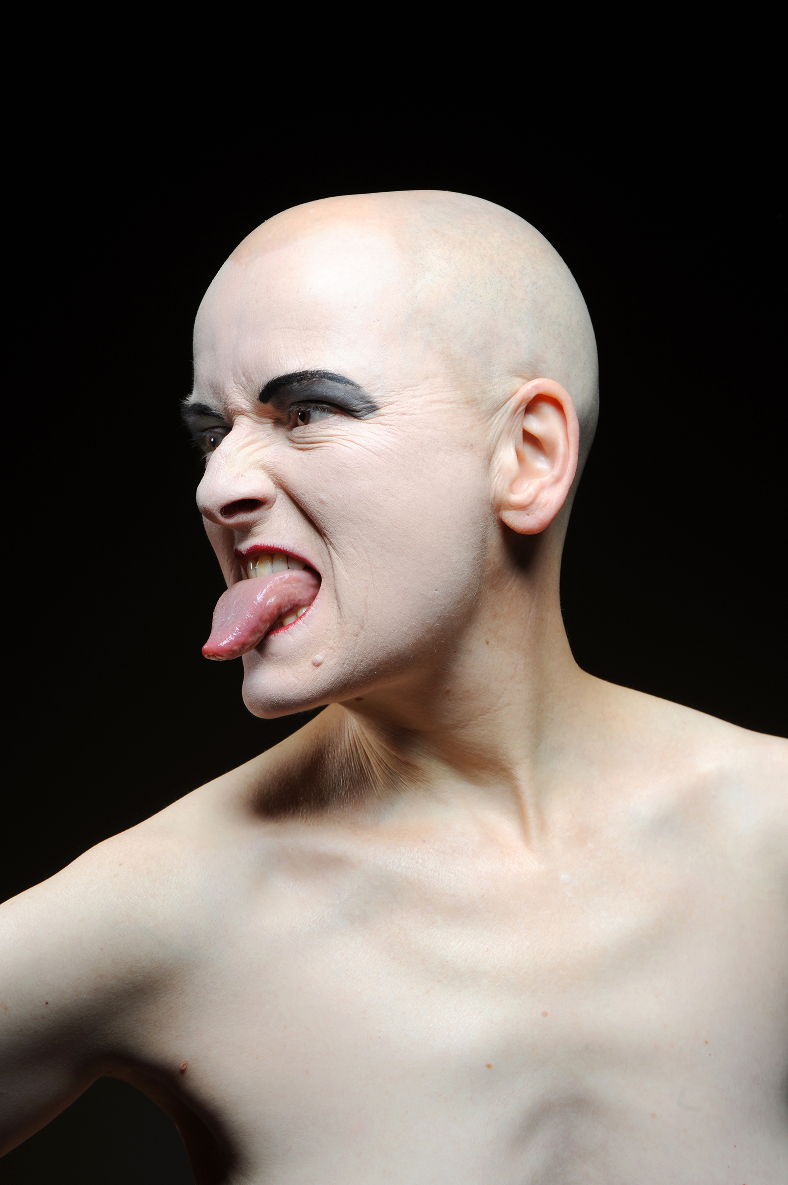 A person with a bald head and make-up sticks out his tongue.