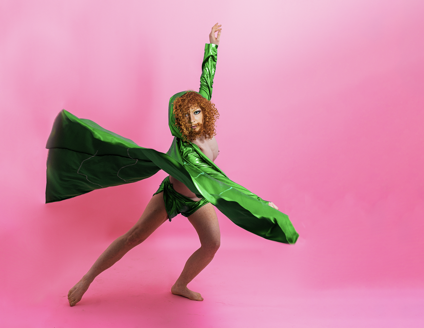 A person with red curls, a full beard and a bright green cape dances in front of a pink background.