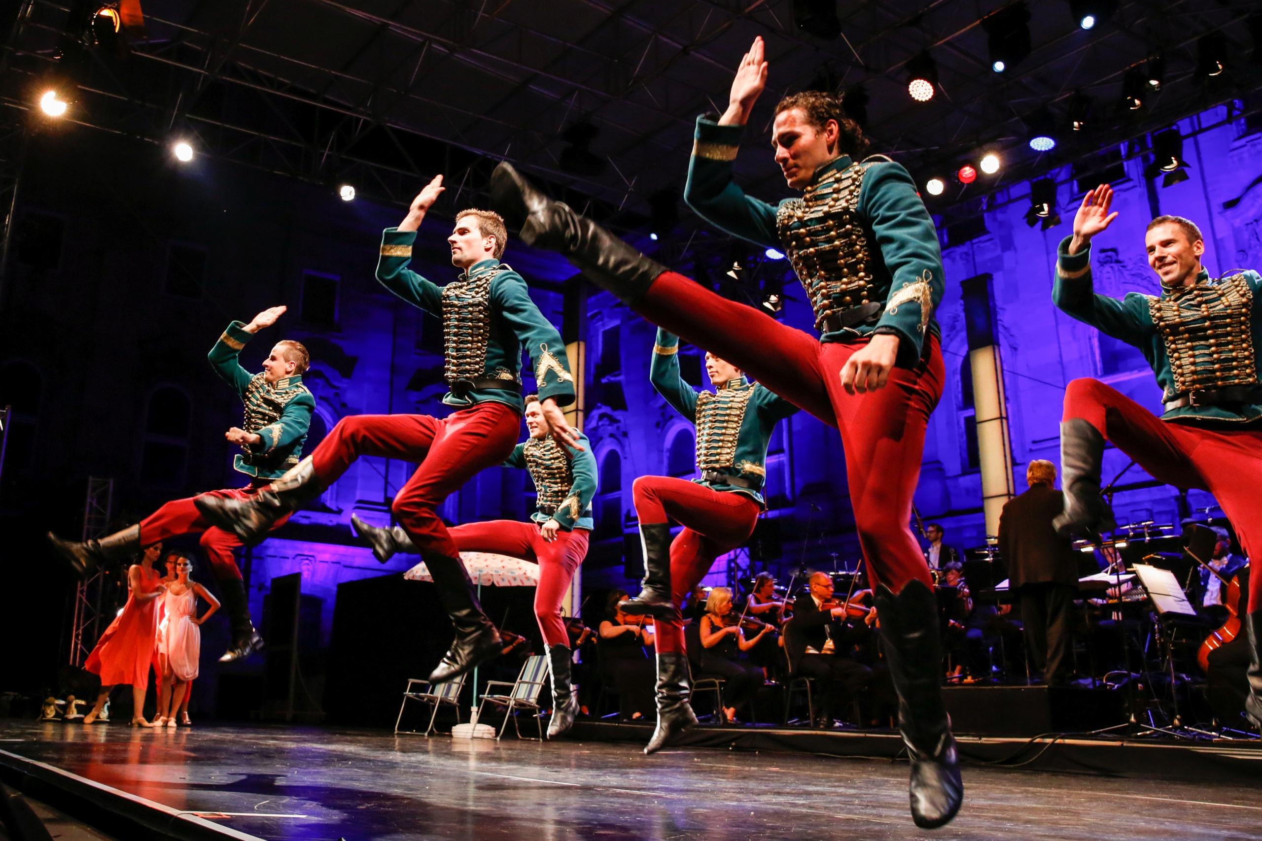 Several men in red pants and gold-trimmed green jackets jump in dance. An orchestra sits in the background.